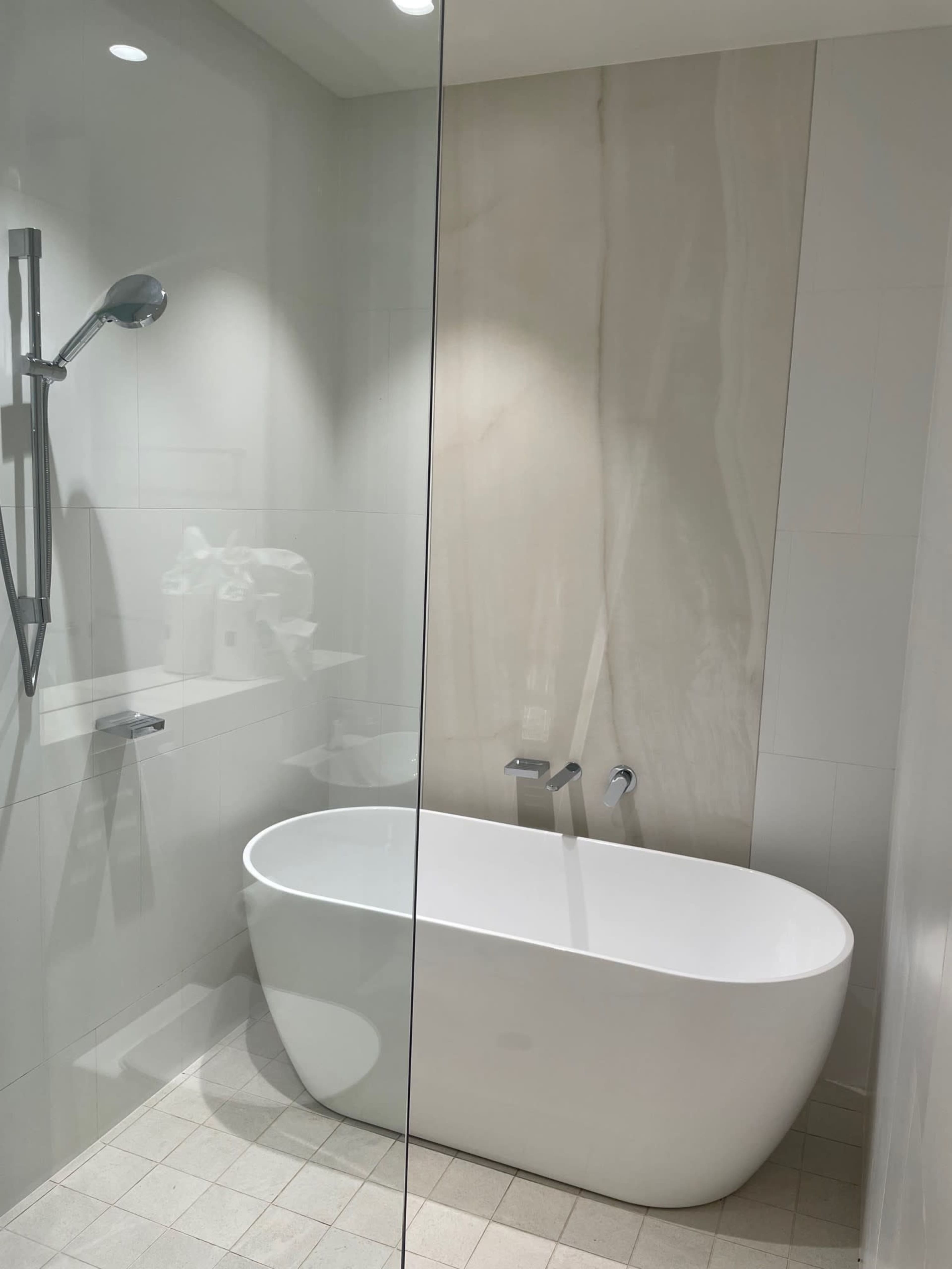 The image shows a modern bathroom featuring a freestanding white bathtub, a shower area with a glass partition, and light-colored tiled walls.