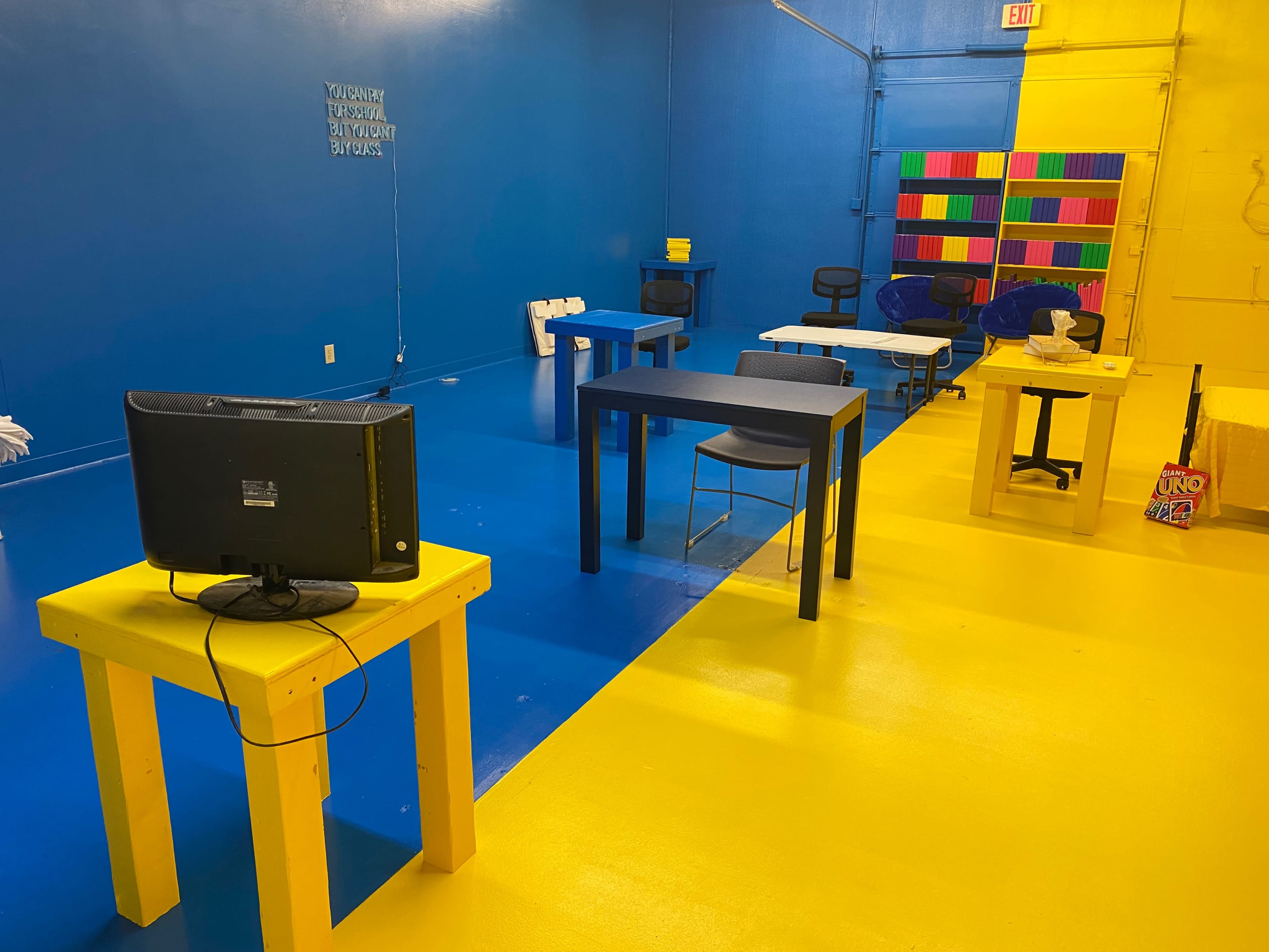 The image shows a brightly colored room with blue and yellow floors, featuring several tables and chairs, along with an old television set on a yellow table.