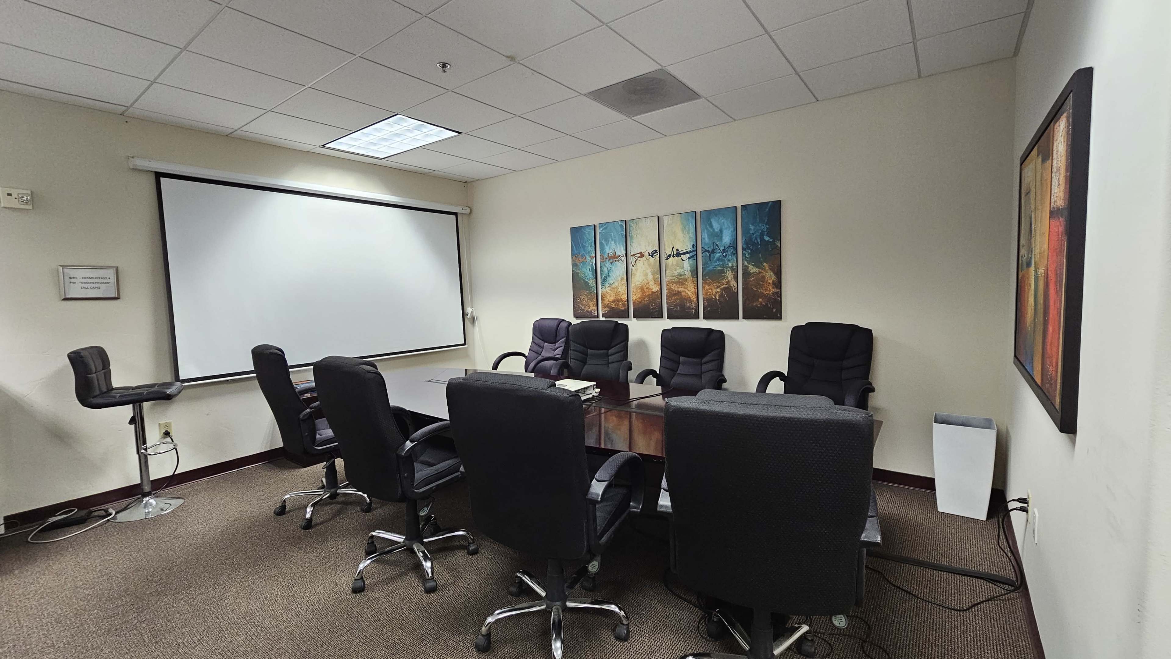 A conference room features a large table surrounded by several black office chairs, with a projector screen and artworks on the walls.