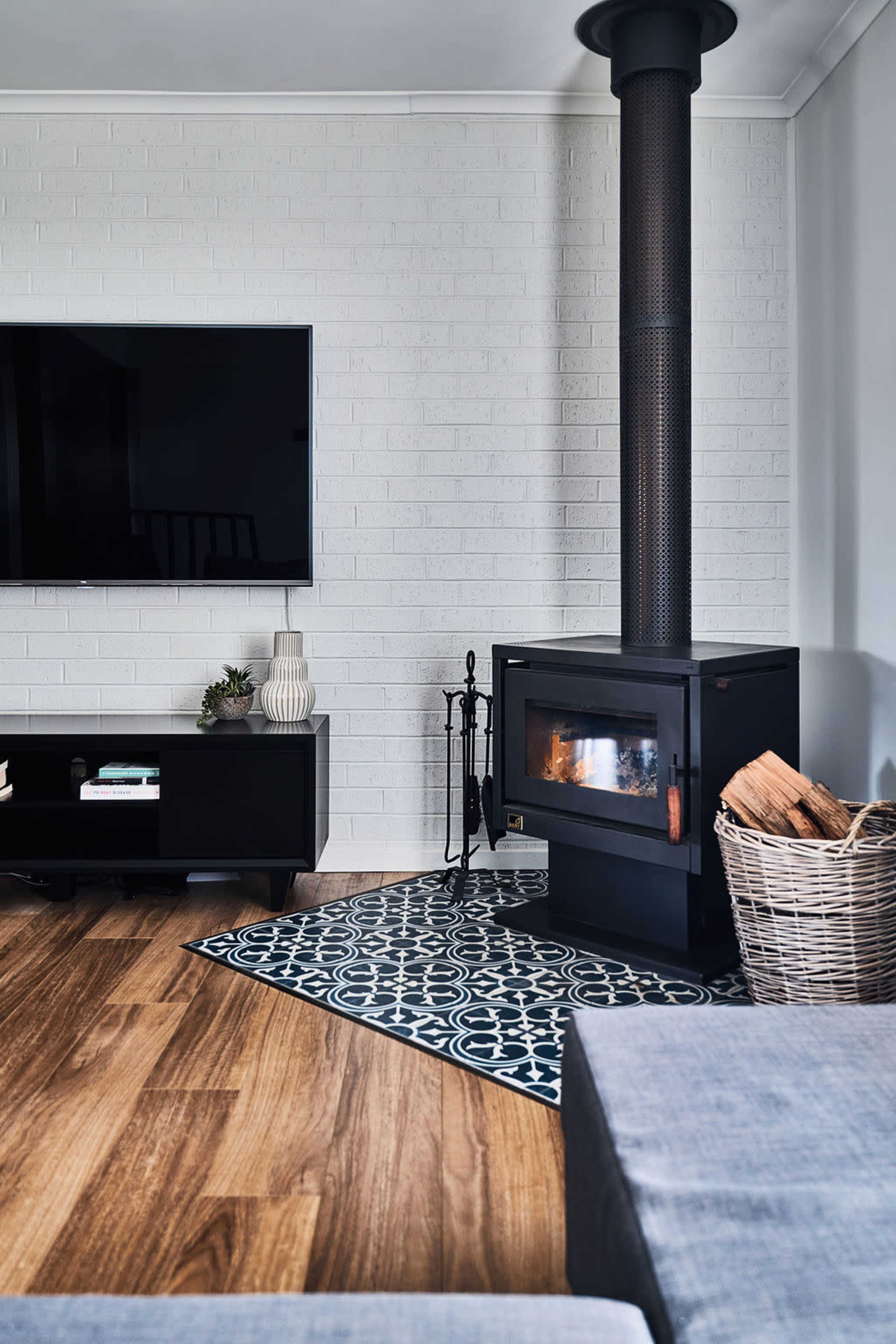 A modern living room featuring a black wood stove, a flat-screen TV, a low black television unit, and a woven basket beside a patterned area rug on a wooden floor.