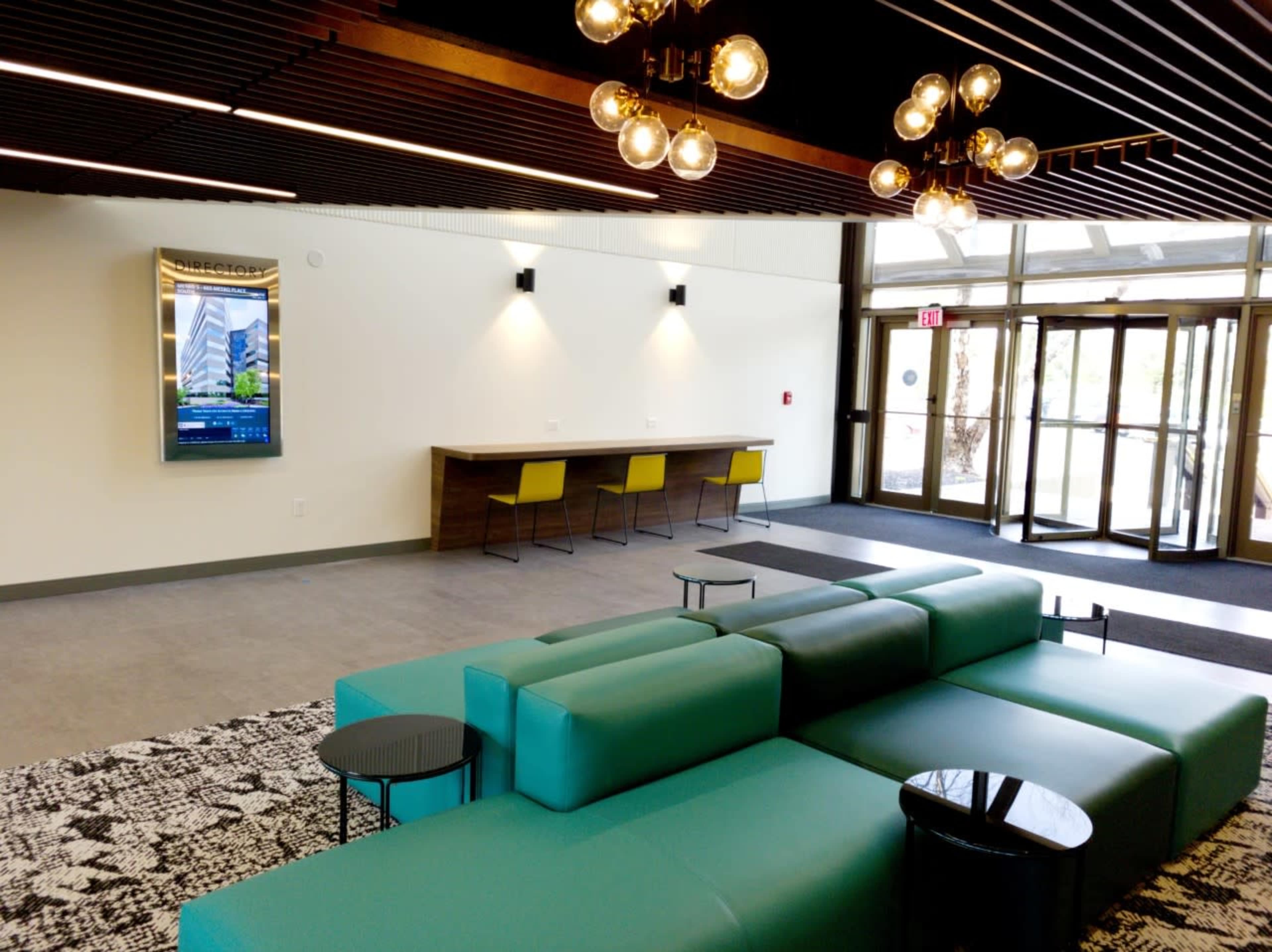 A modern lobby with a green sectional sofa, a low table, a digital display, and a counter with yellow chairs near a large glass entrance.
