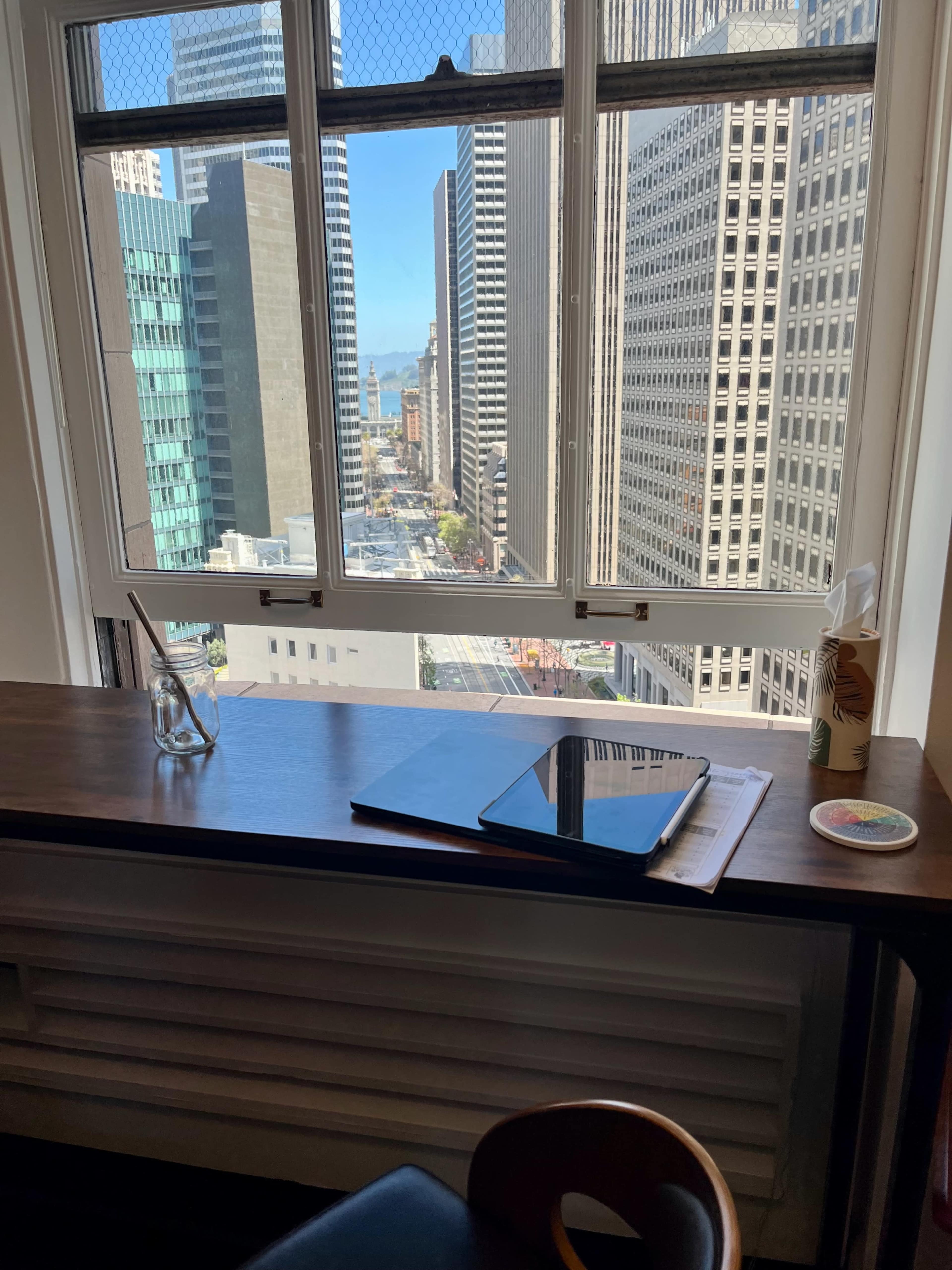 A workspace with a notebook and pen sits adjacent to a large window overlooking a cityscape with tall buildings and a street below.