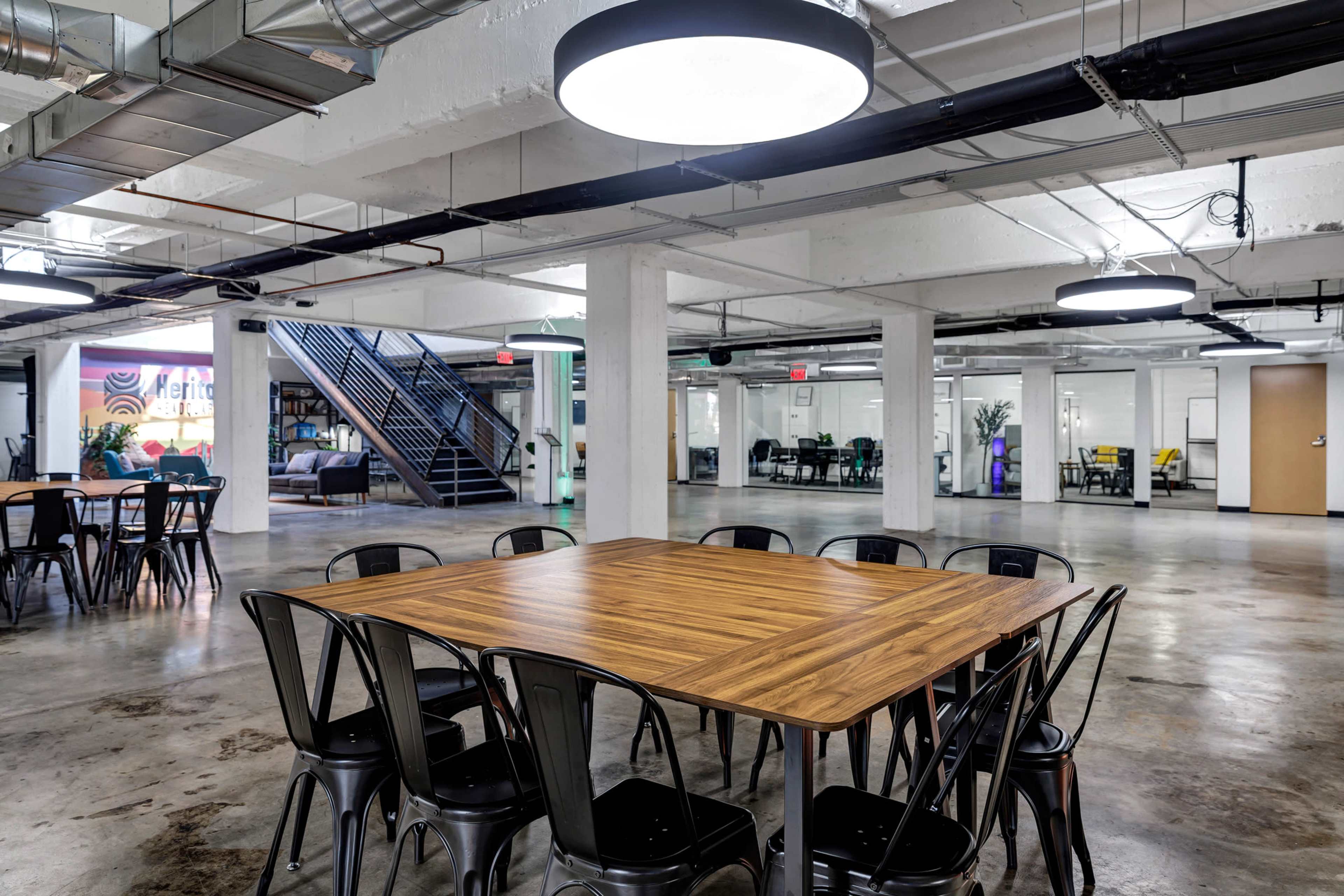 The image shows an industrial-style office space with a large wooden table surrounded by black chairs, visible lighting fixtures, and an open layout featuring multiple work areas in the background.