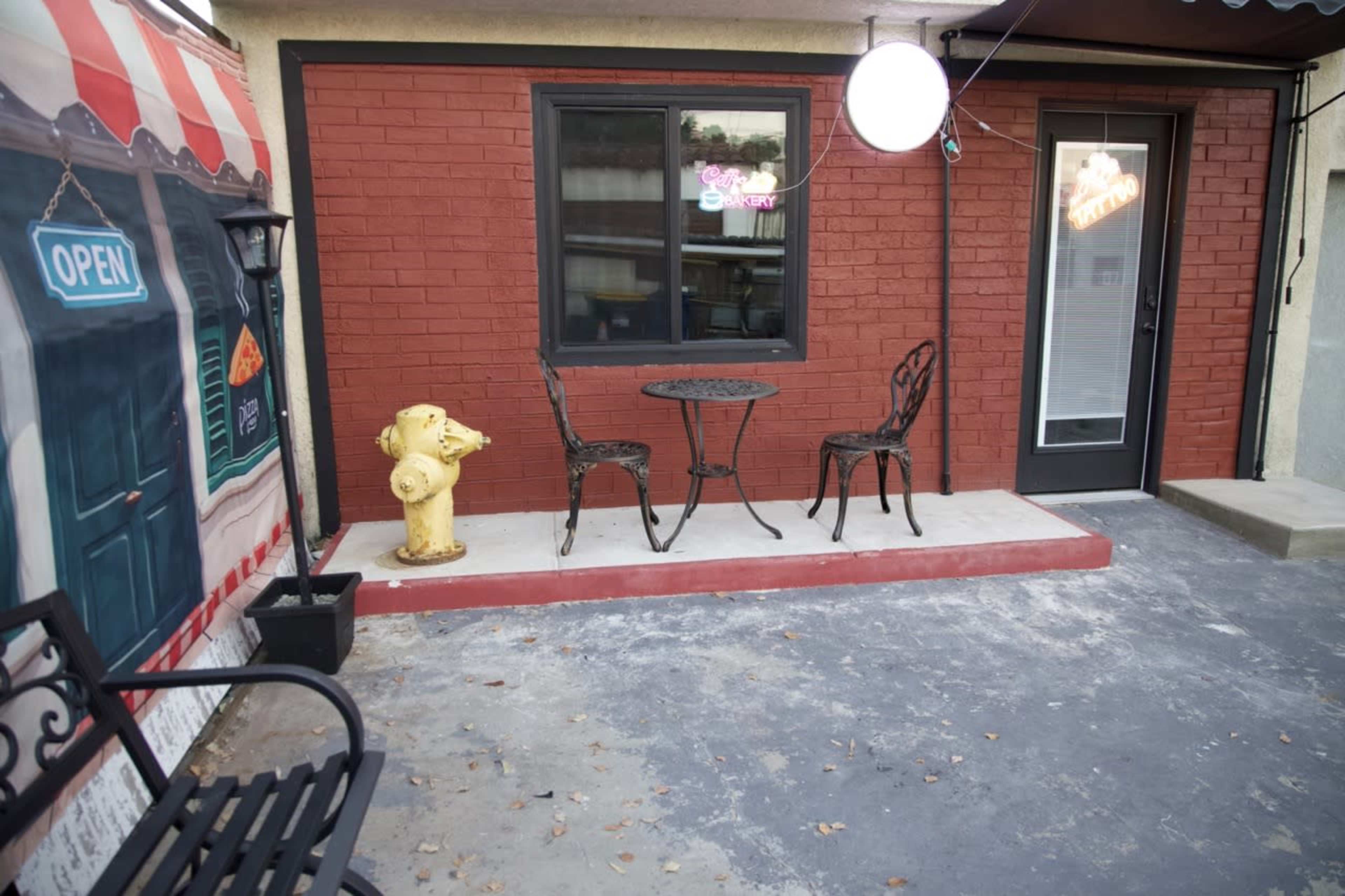 The image shows a small outdoor seating area with a round table and two chairs next to a yellow fire hydrant, against a backdrop of a brick wall and a neon "open" sign.