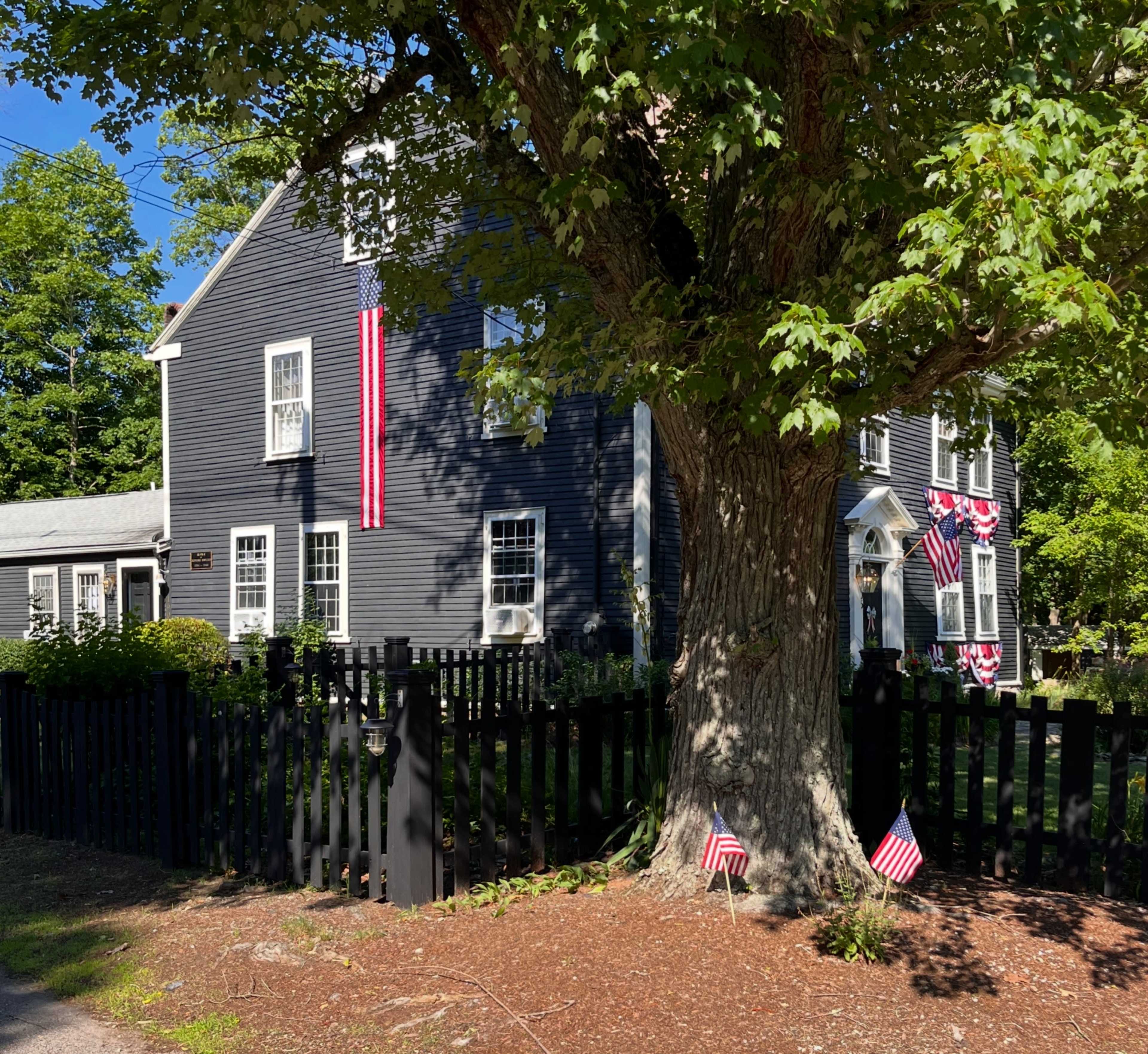 A gray house with white trim displays American flags and is surrounded by a black wooden fence and a large tree.