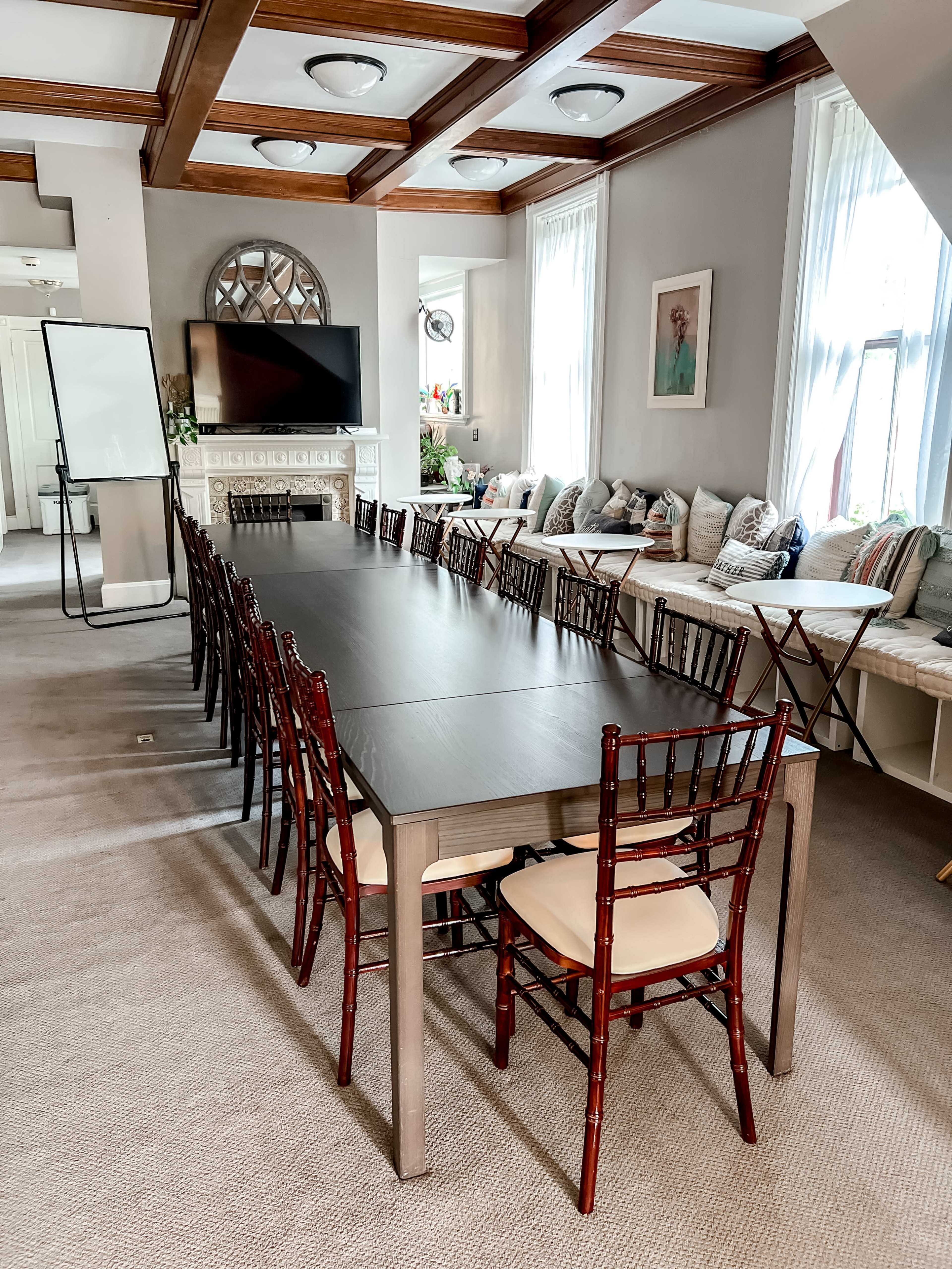 A long conference table is set up in a well-lit room with a fireplace, surrounded by chairs and featuring large windows with decorative cushions nearby.