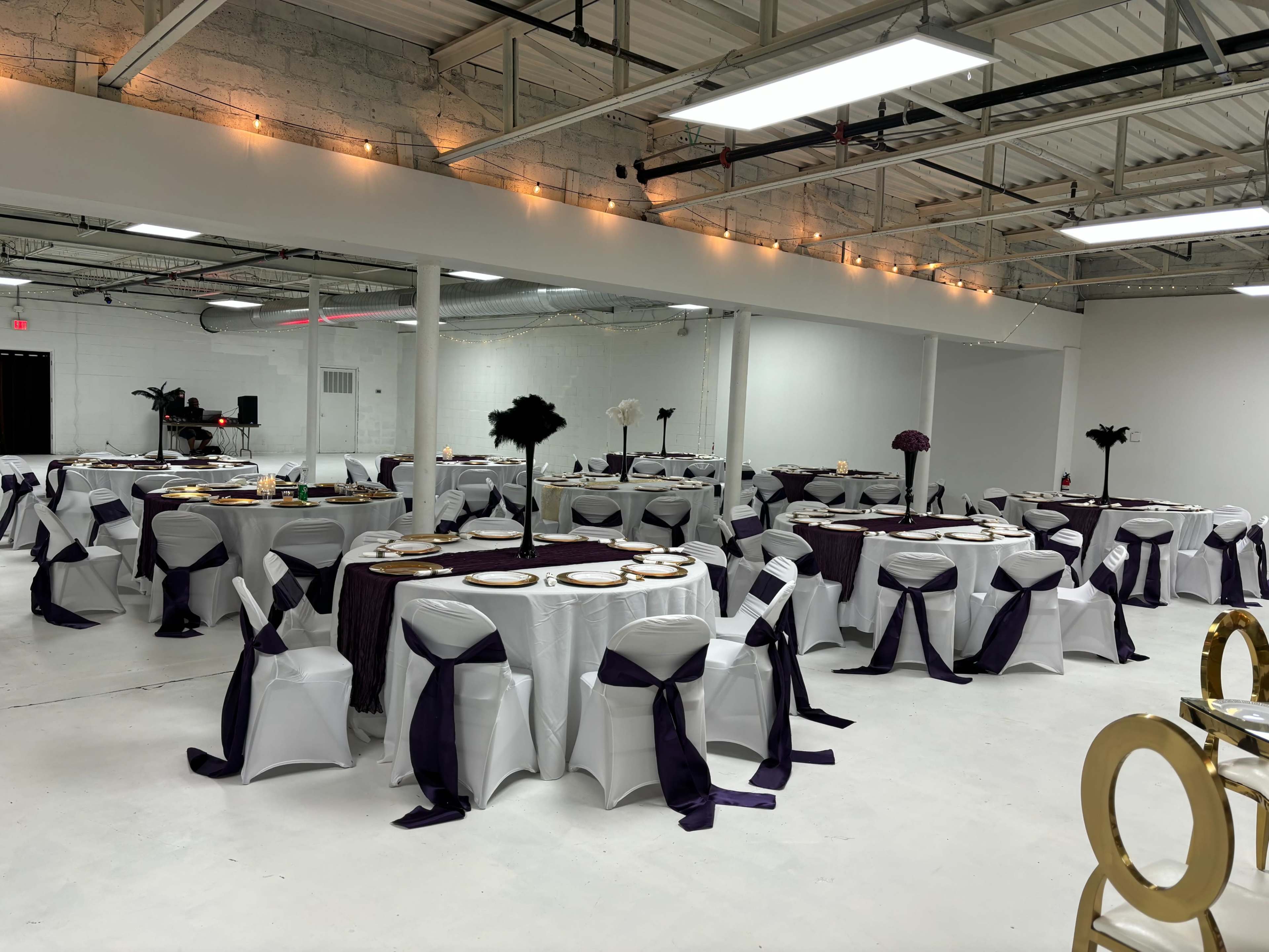 A large event space features several round tables set with white tablecloths and dark purple accents, surrounded by white chairs adorned with purple sashes.