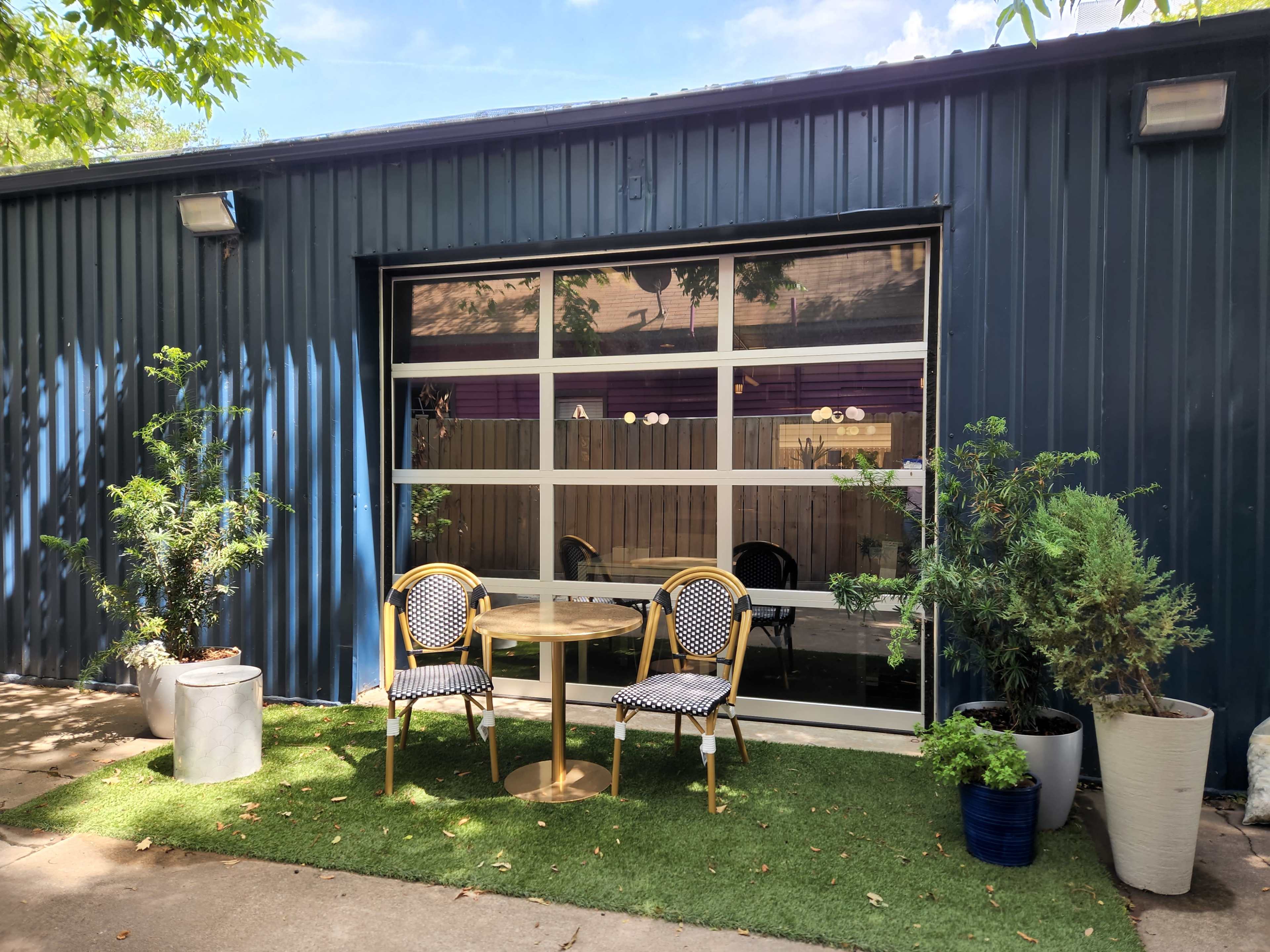 A small outdoor seating area with two chairs and a table in front of a large window on a blue metal building, surrounded by potted plants.