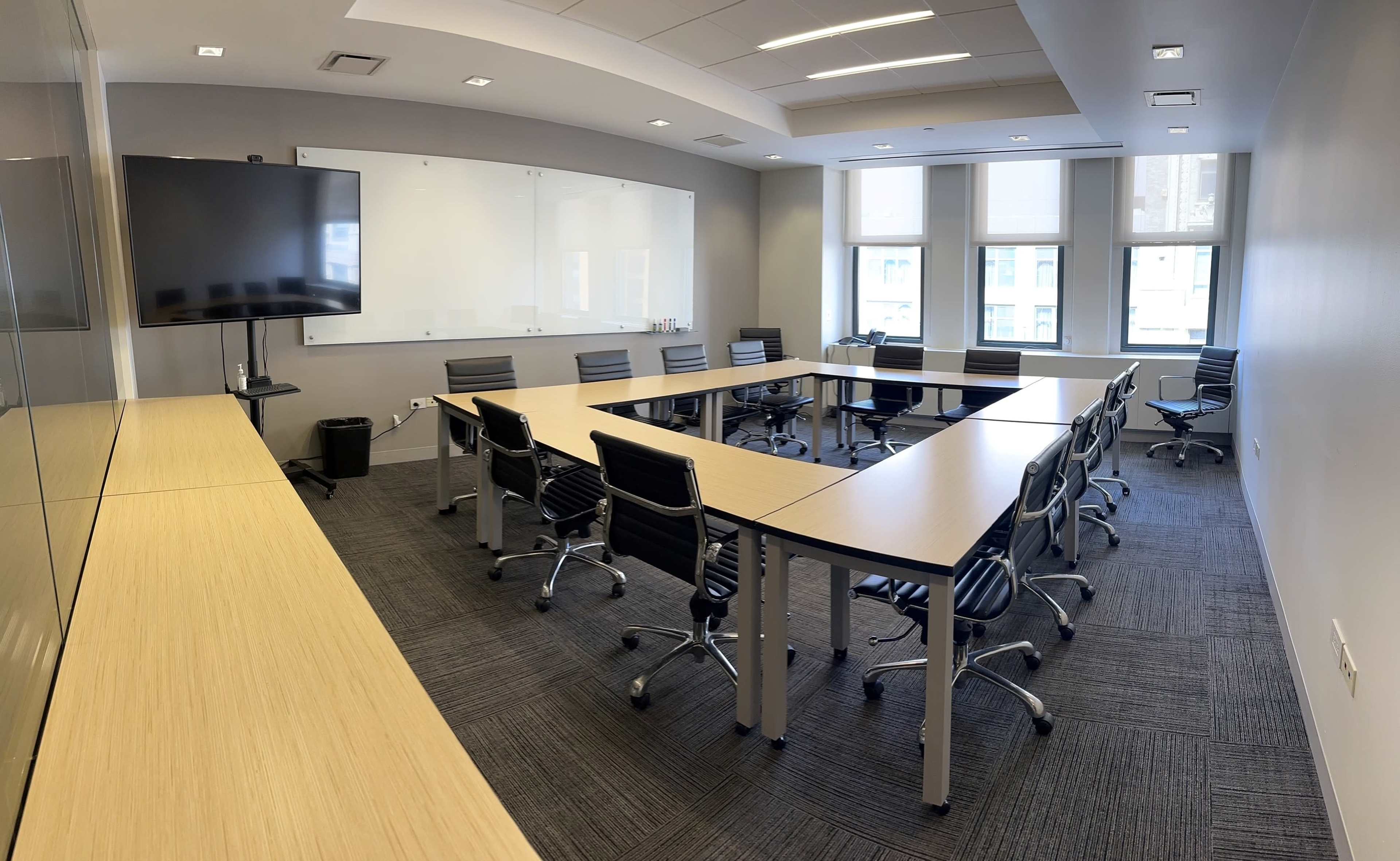 A modern conference room features a large rectangular table surrounded by ergonomic chairs, with a wall-mounted screen and windows offering natural light.