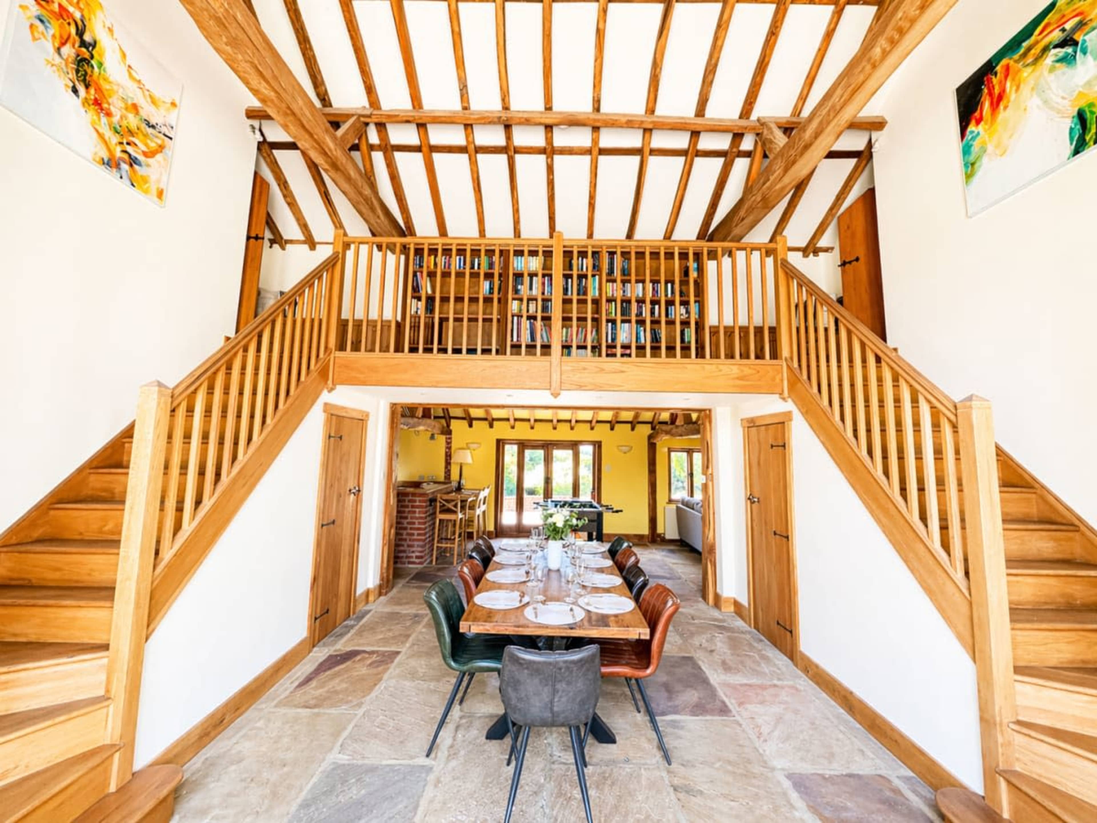 The image shows a spacious interior featuring a wooden staircase leading to a loft area, with a dining table set in the center and large windows illuminating the room.