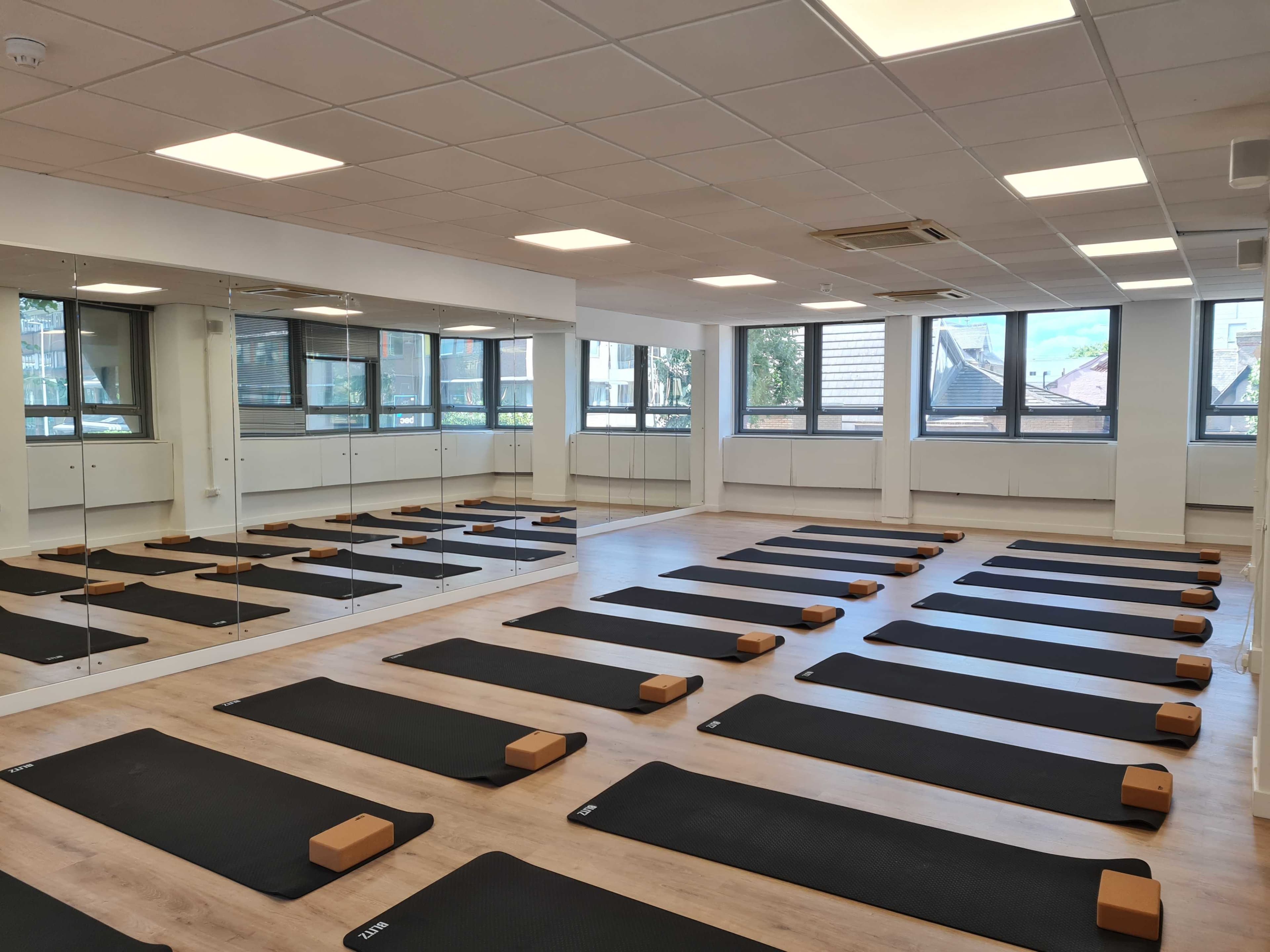 The image shows a spacious yoga studio with several black mats lined up on the floor and wooden blocks placed at the front of each mat, alongside large mirrors and windows.