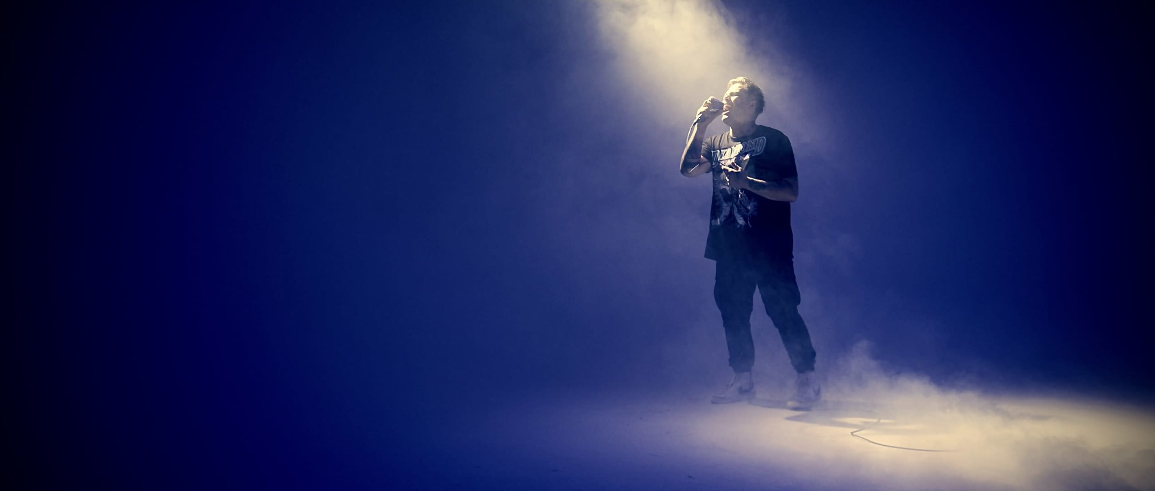 A singer stands in a spotlight, surrounded by a haze of smoke on a dark stage.