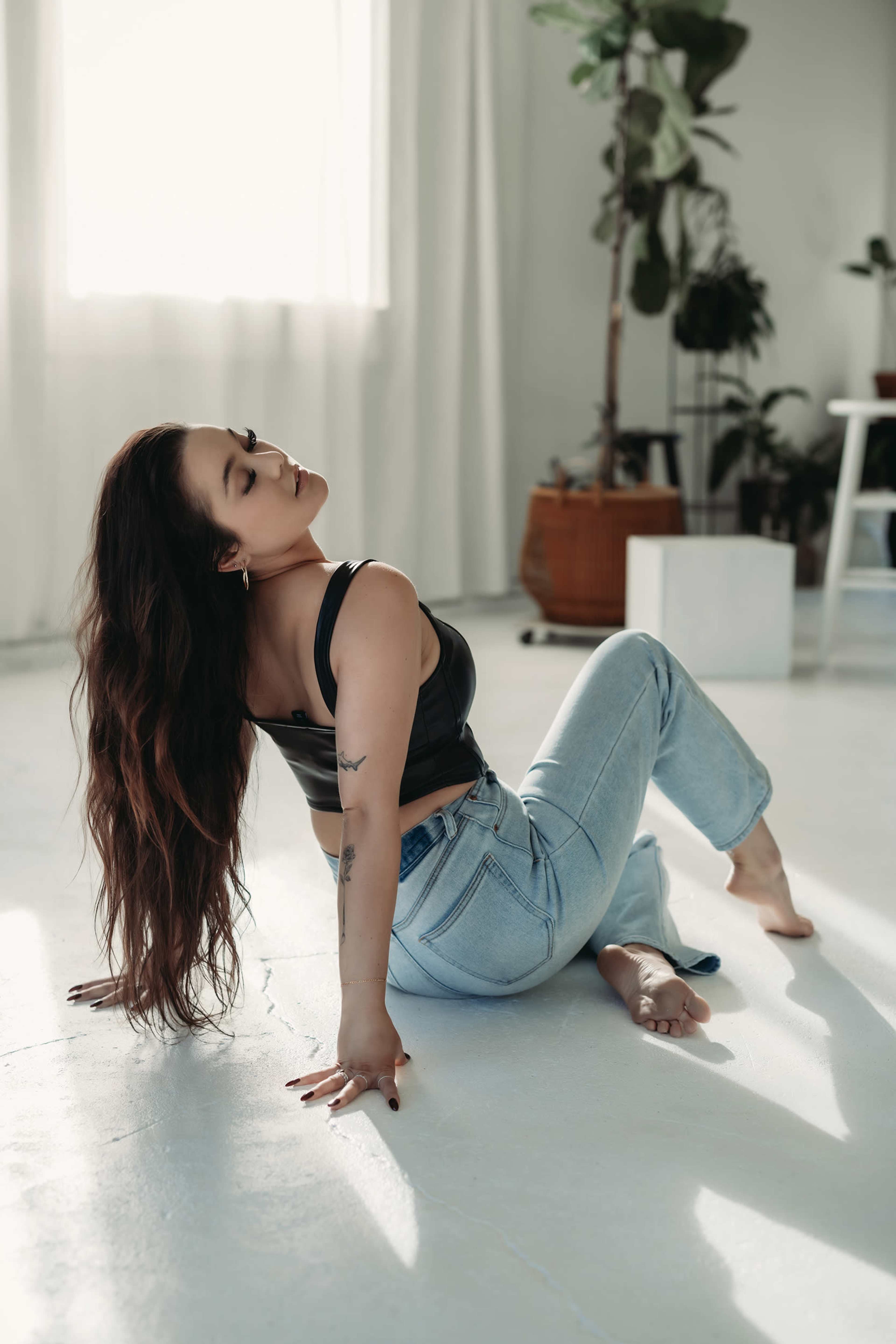 A woman in a black tank top and light blue jeans poses gracefully on the floor in a well-lit room filled with plants.
