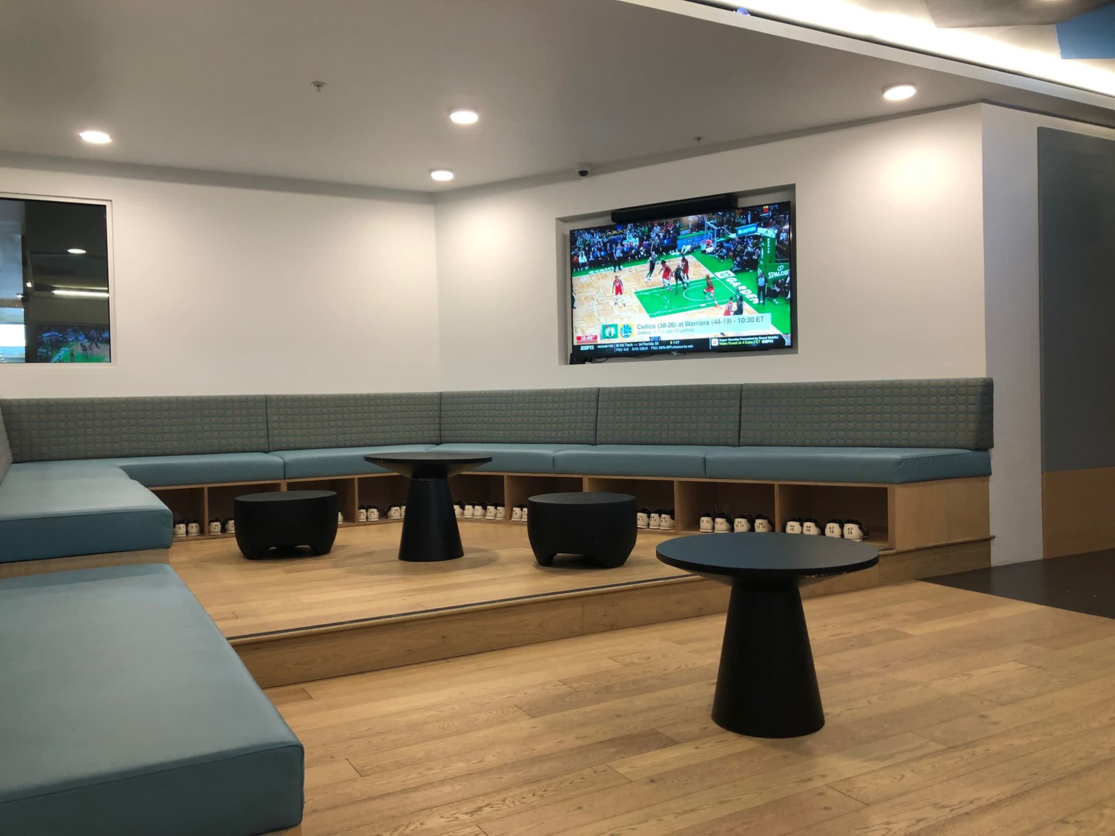 A modern lounge area with wooden flooring, a series of low couches arranged in a circular layout, and a television displaying a basketball game on the wall.