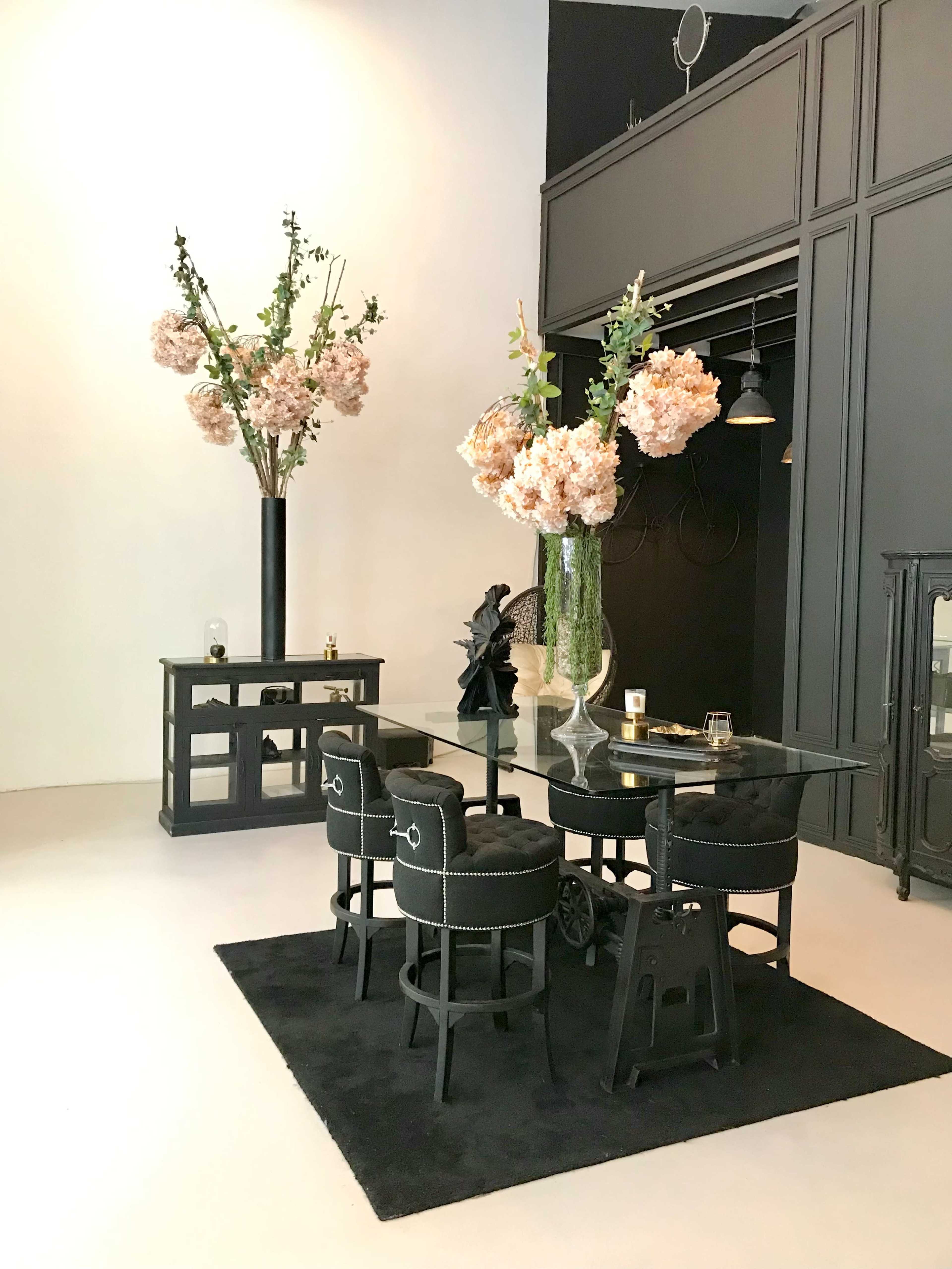 A modern dining area features a glass table surrounded by four black upholstered stools, adorned with tall floral arrangements in stylish vases.