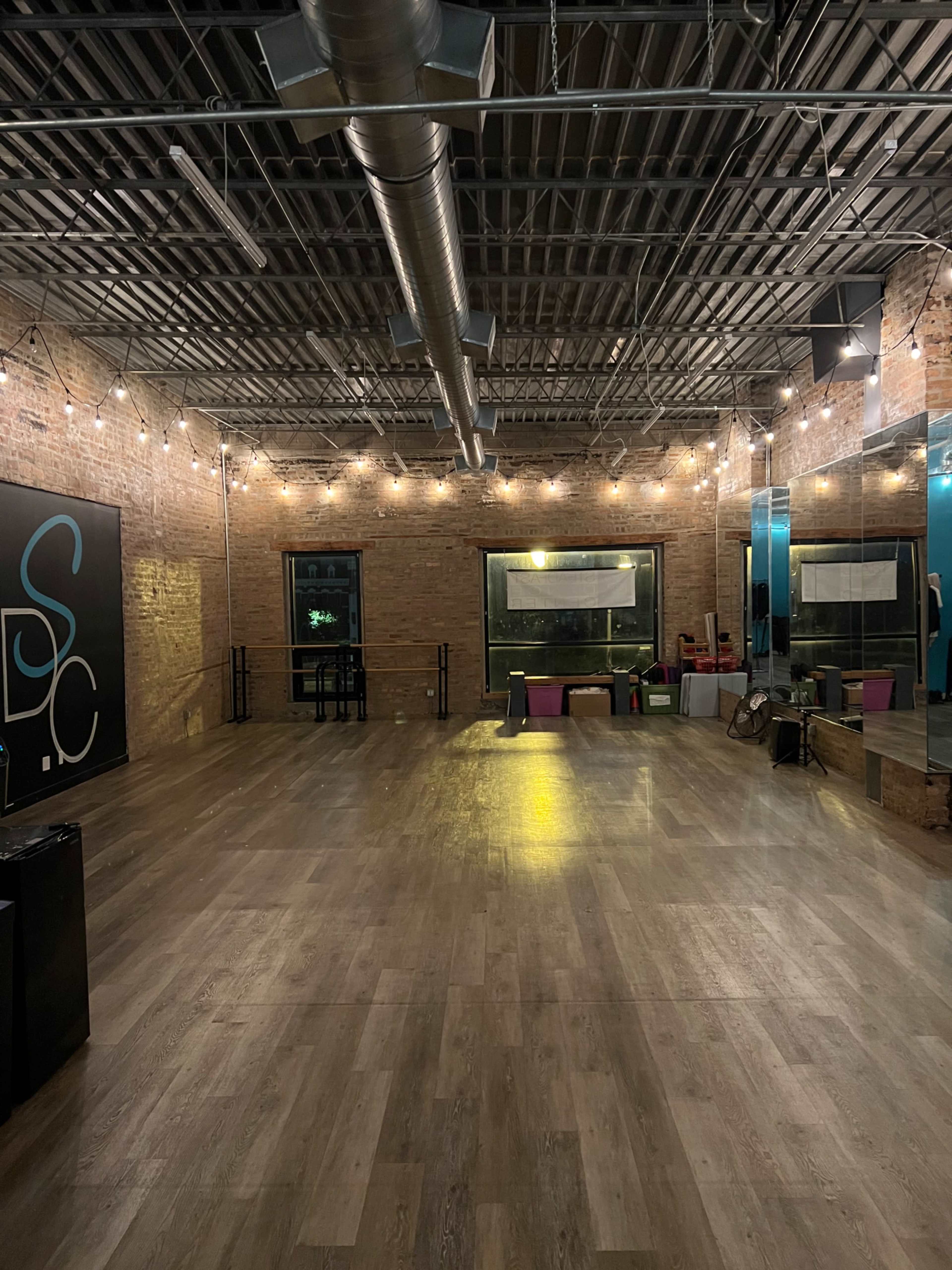 The image shows an empty dance studio with wooden flooring, exposed brick walls, and large mirrors, illuminated by string lights.