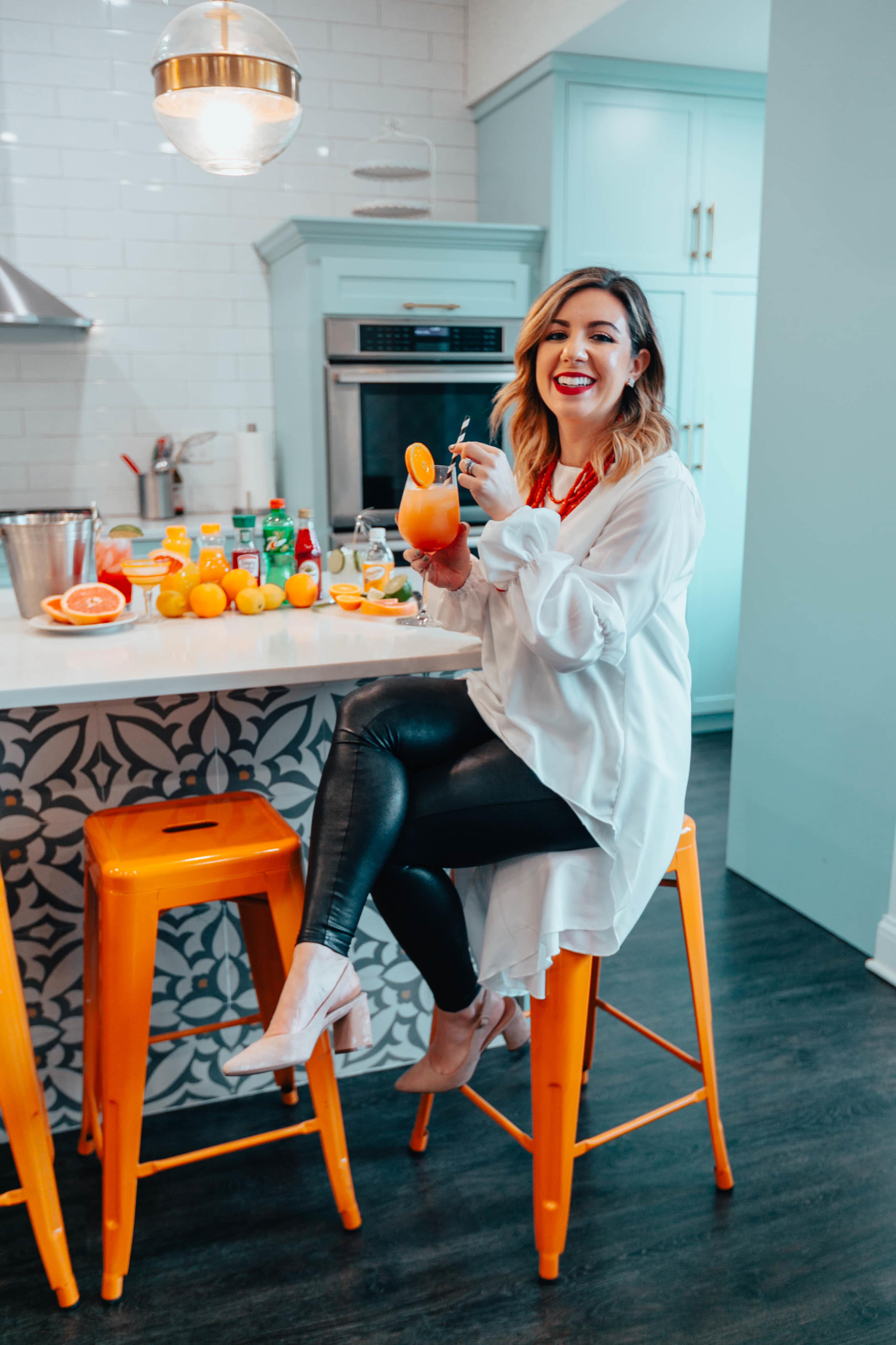 A person sits on an orange bar stool at a kitchen island, holding a beverage and surrounded by various fruits and ingredients.