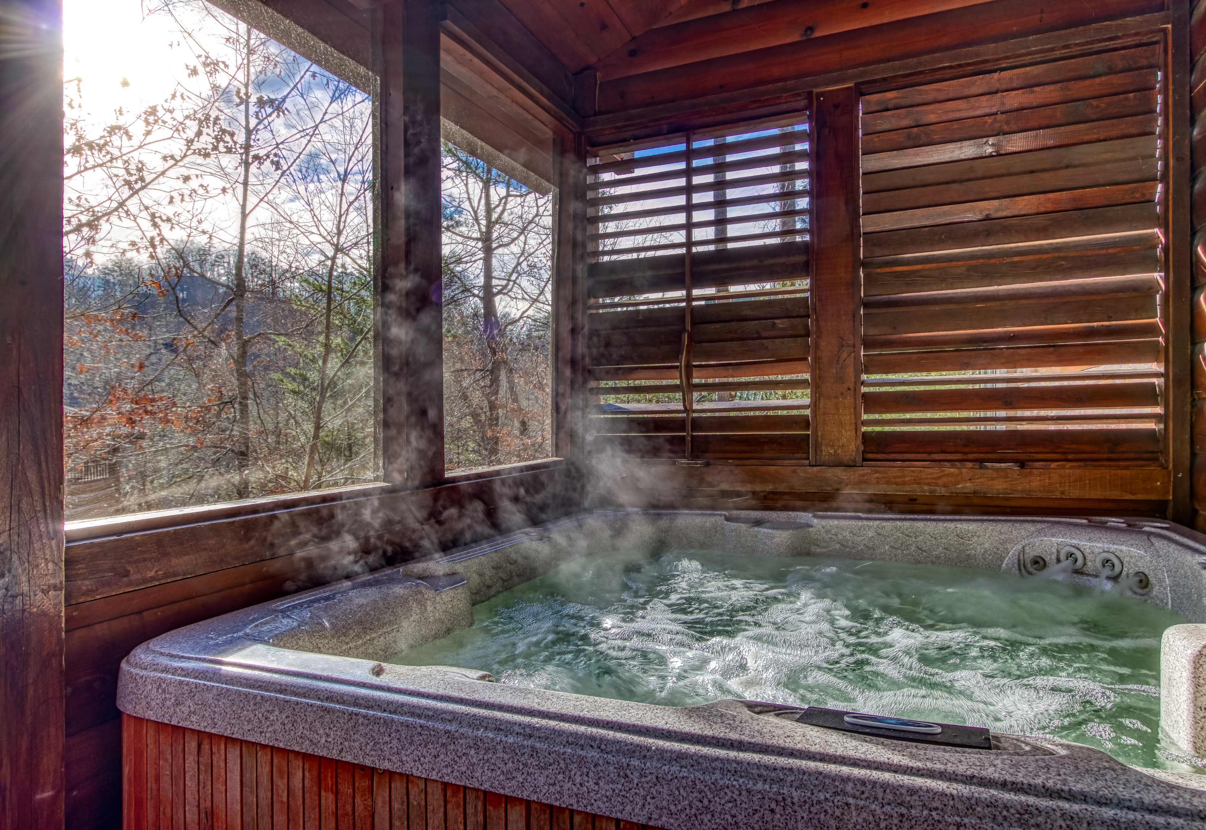 A hot tub with steam rising is located inside a wooden structure with slatted windows, surrounded by trees.