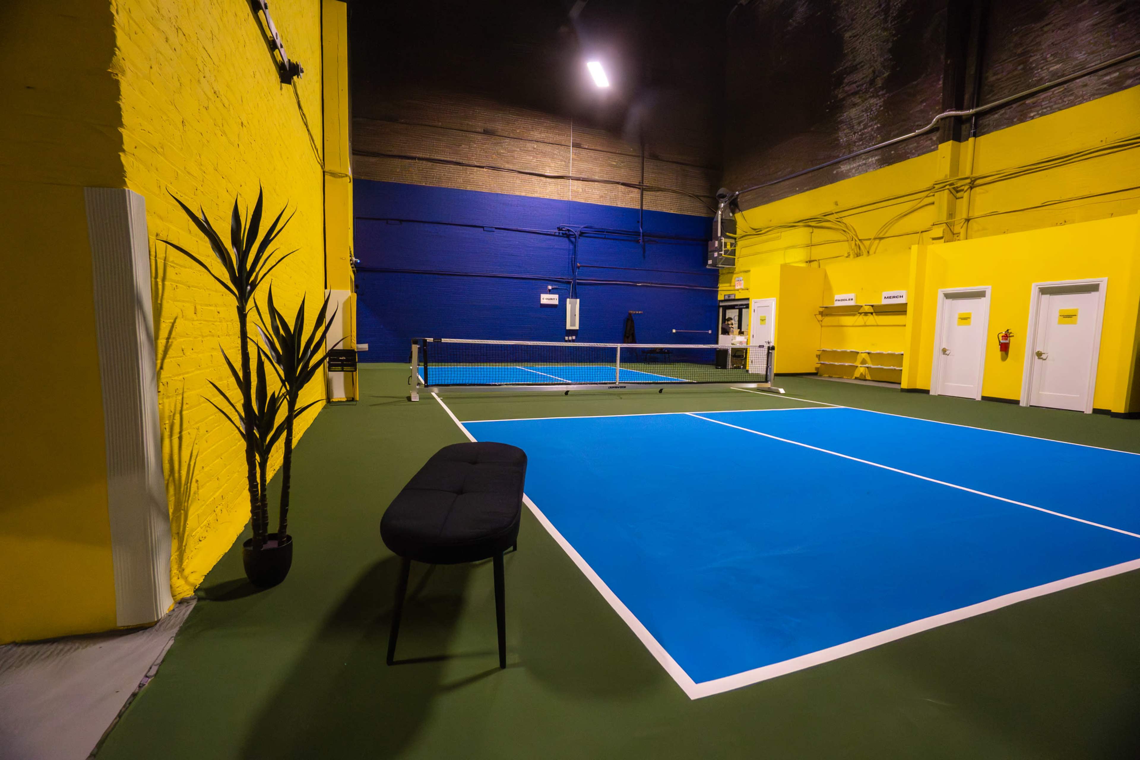 The image shows a brightly colored indoor tennis court with blue and yellow walls, featuring a net and a bench.