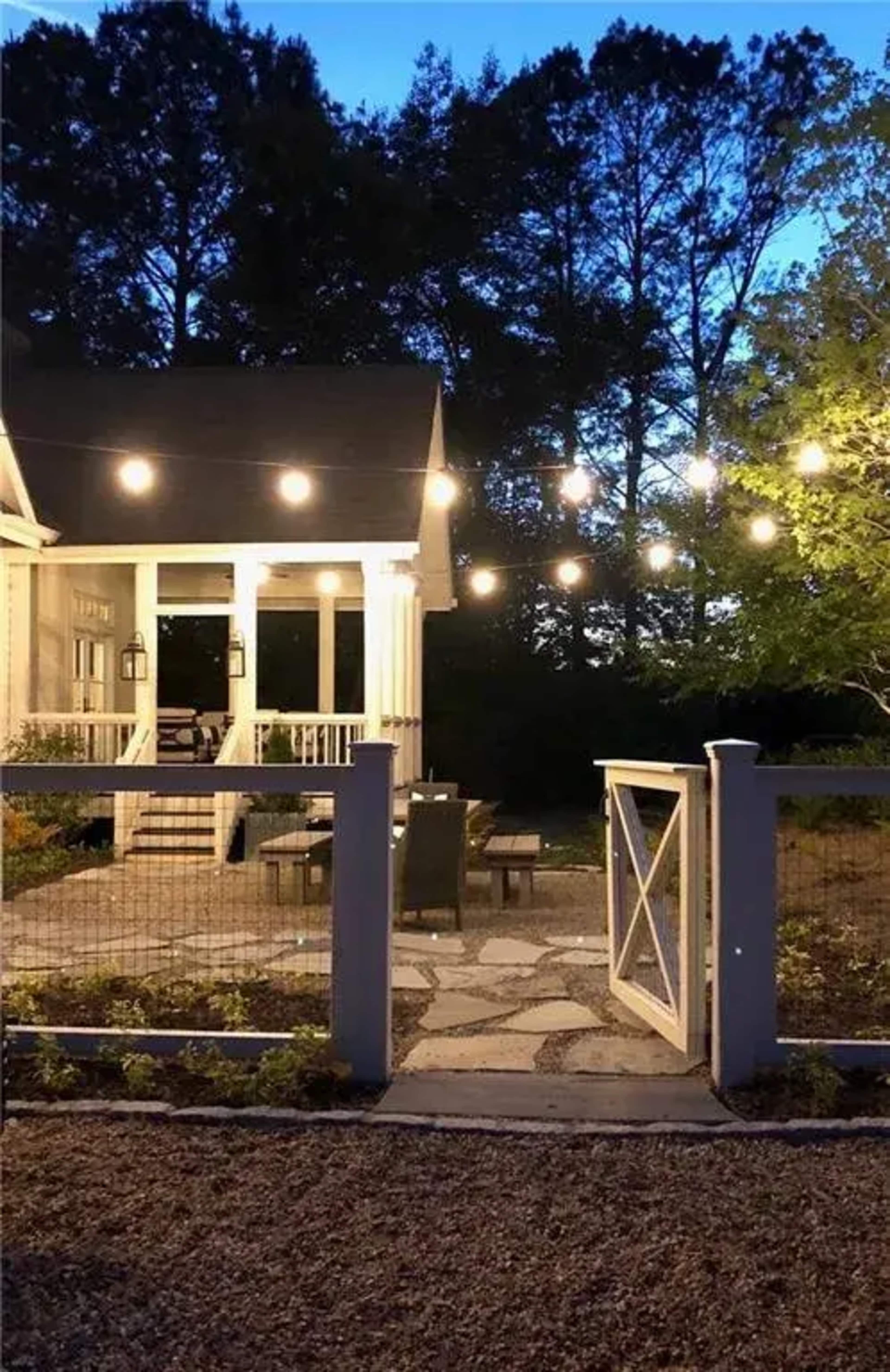 A backyard area with string lights illuminating a seating arrangement on a stone patio, framed by a white gate and surrounded by trees at dusk.