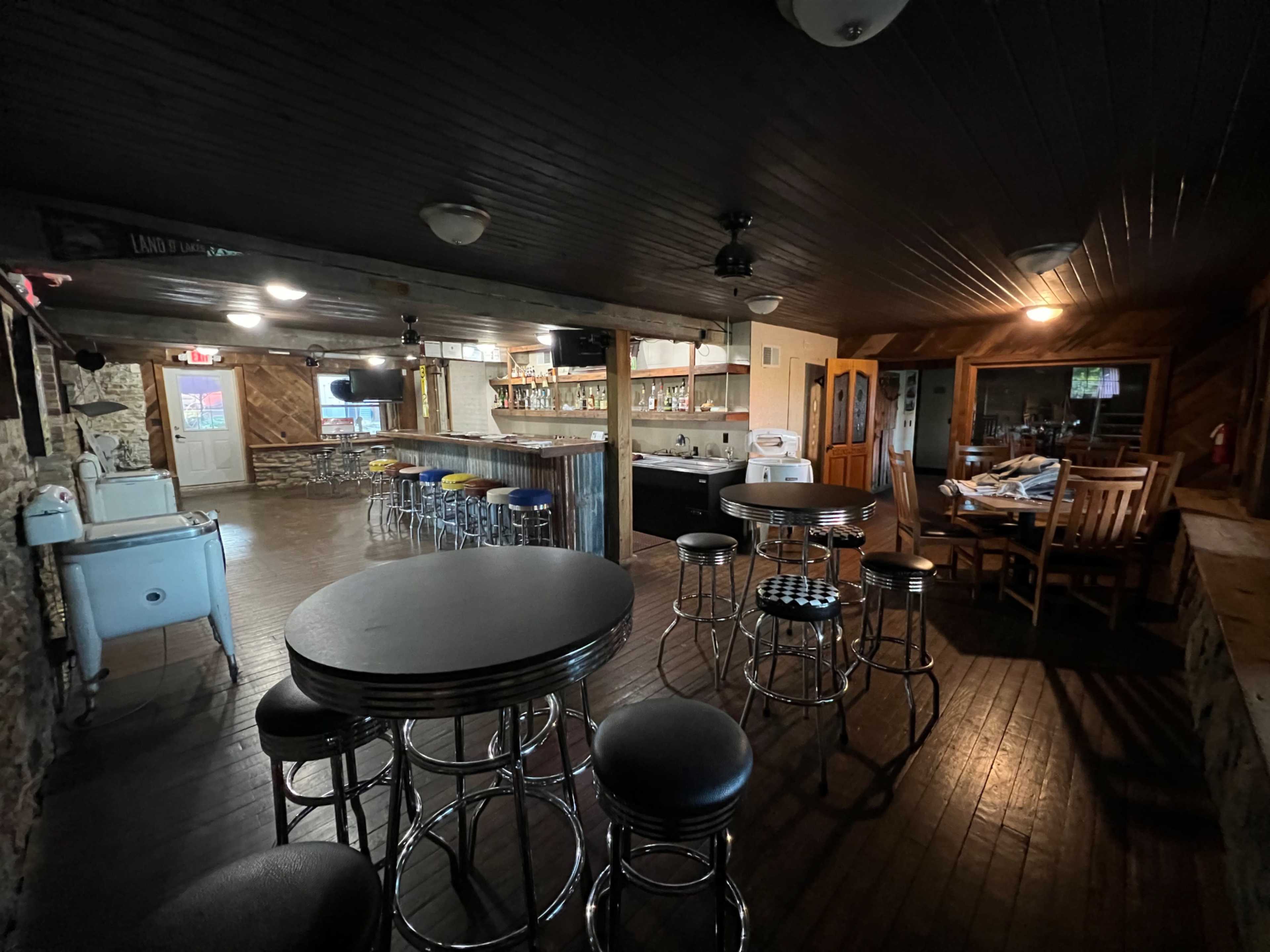 The image shows a rustic bar area with several round tables and stools, a long counter with shelves behind it, and a kitchen area in the background.