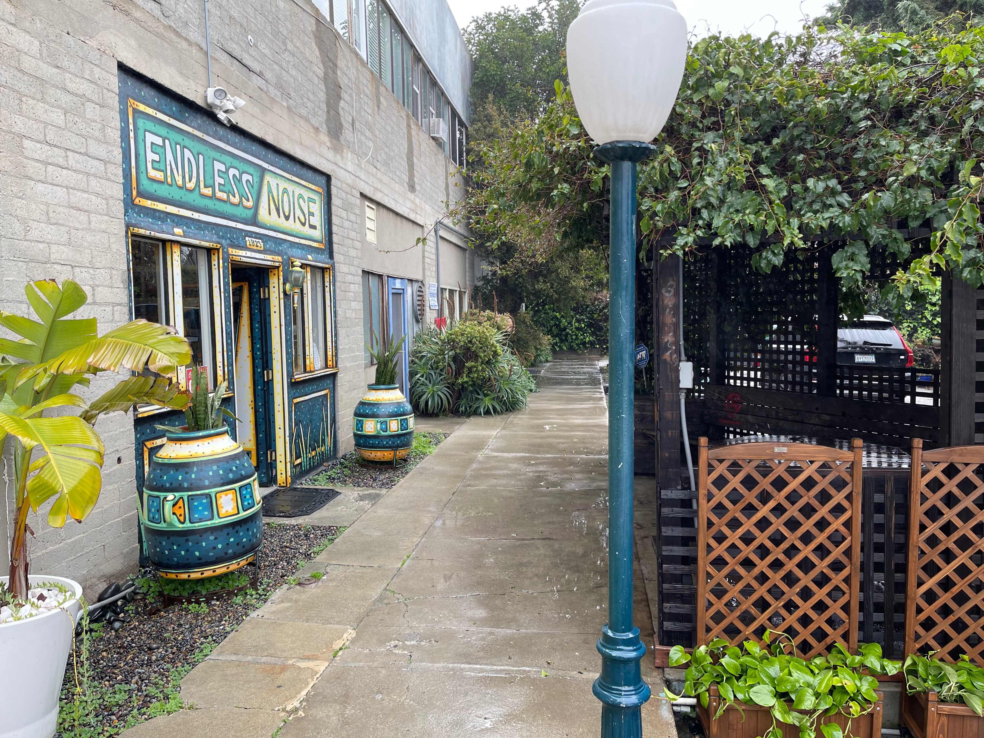 The scene shows a sidewalk beside a building featuring the entrance "ENDLESS NOISE," with decorative barrels and greenery along the path.