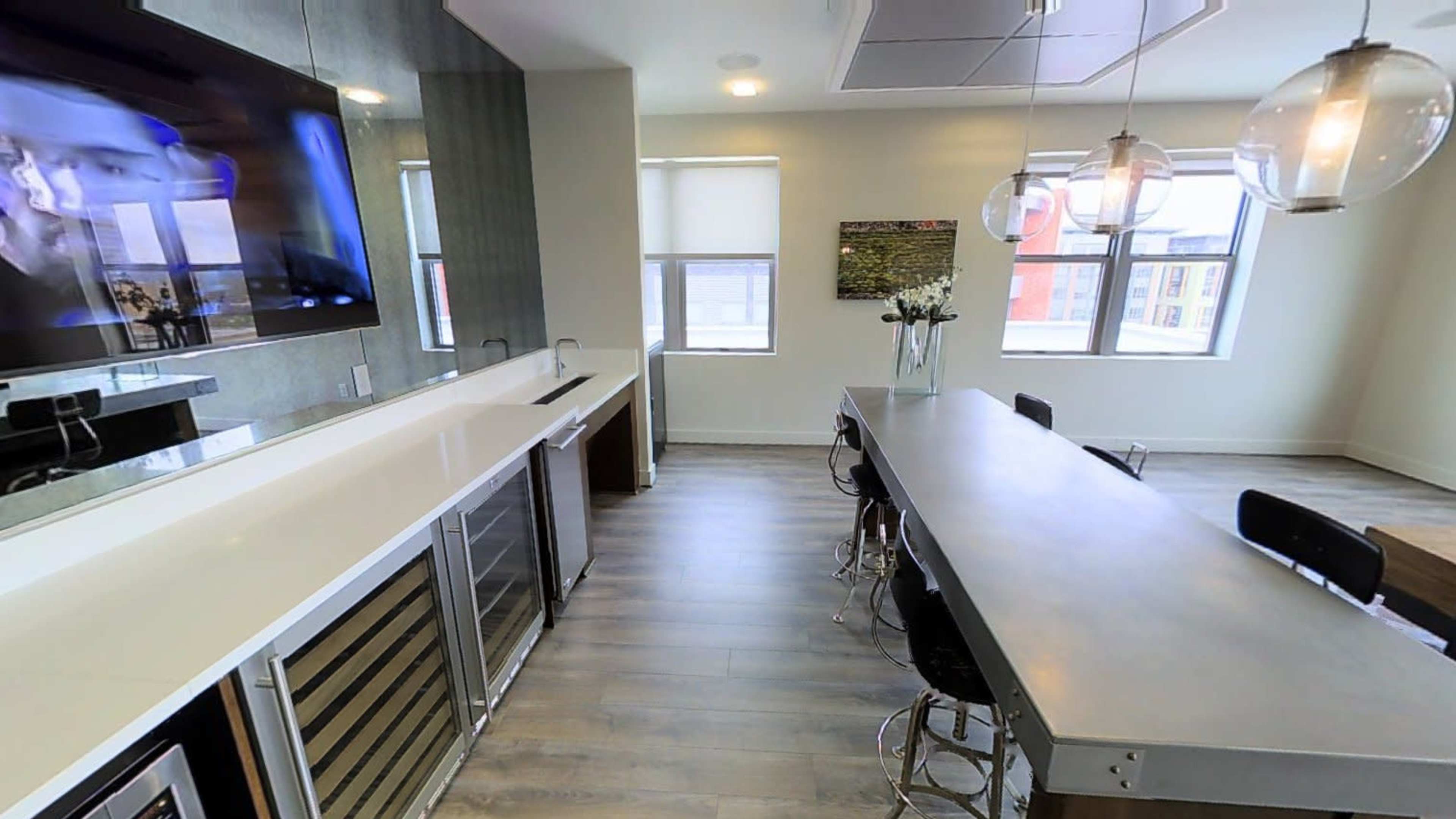 The image shows a modern kitchen with a long dining table, bar stools, and a large wall-mounted television.