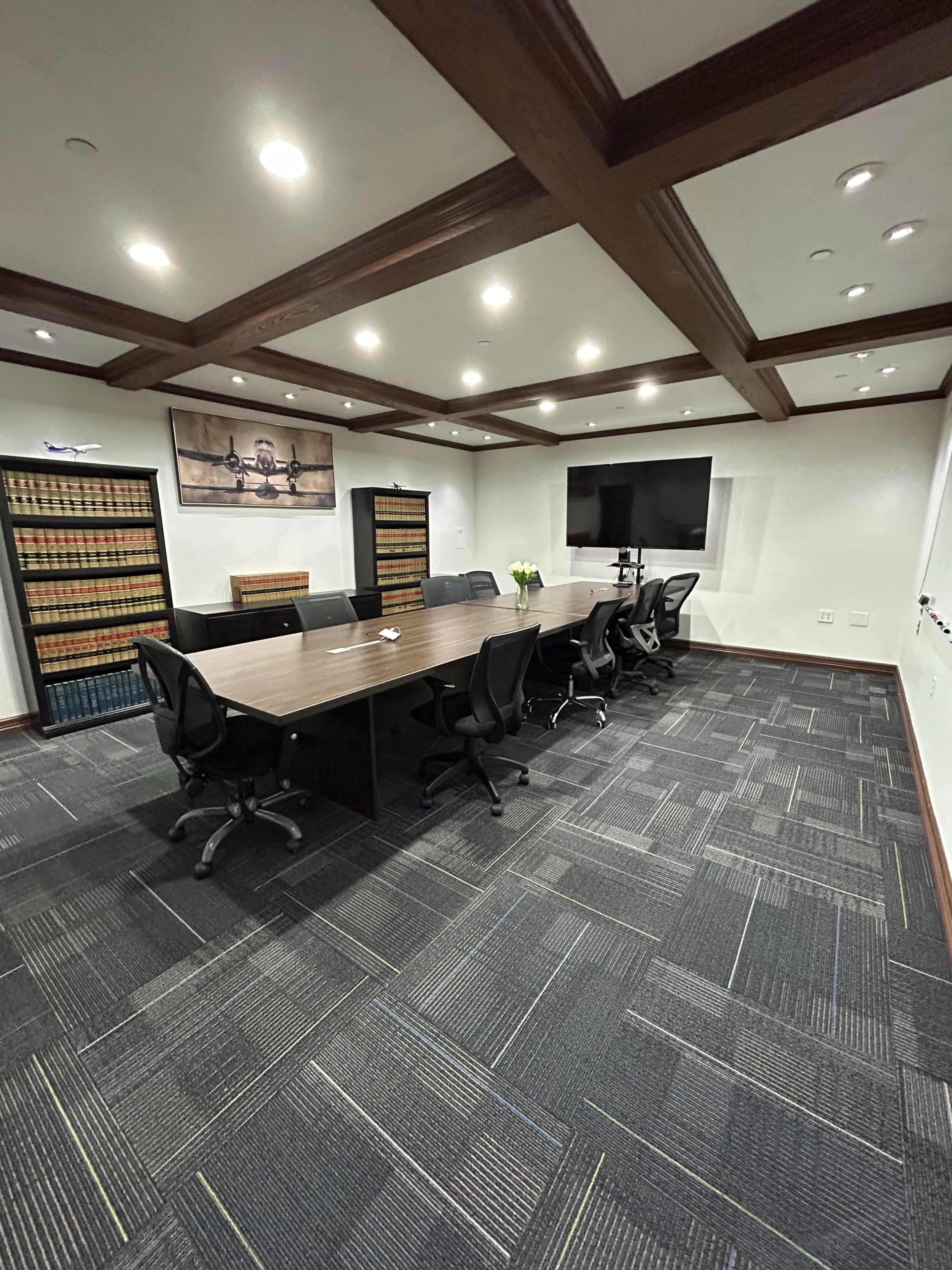 The image shows a modern conference room featuring a long wooden table surrounded by black office chairs, with bookshelves filled with books on one side and a large screen mounted on the wall.