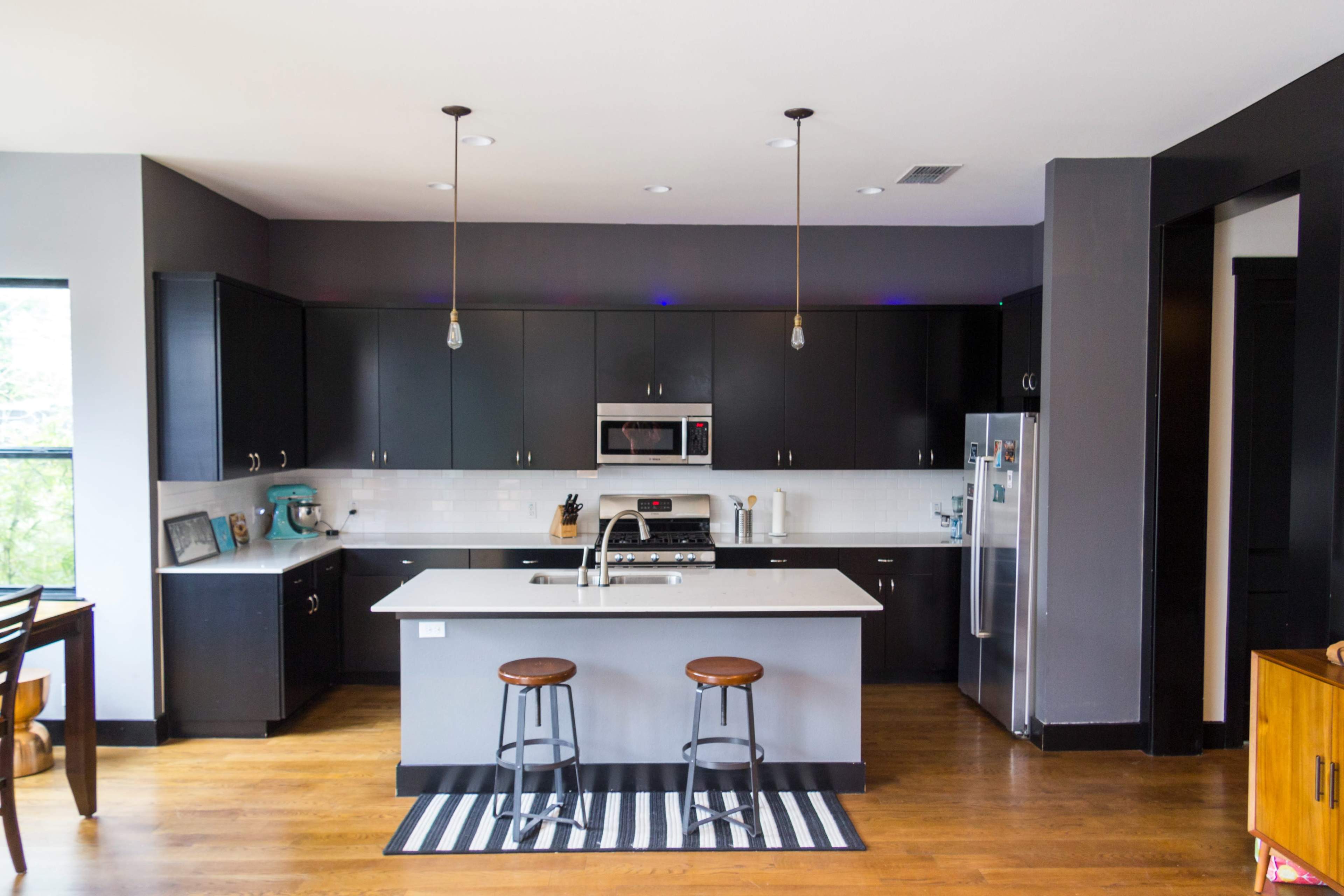 A modern kitchen features dark cabinets, a central island with pendant lighting, and a striped rug on wooden flooring.