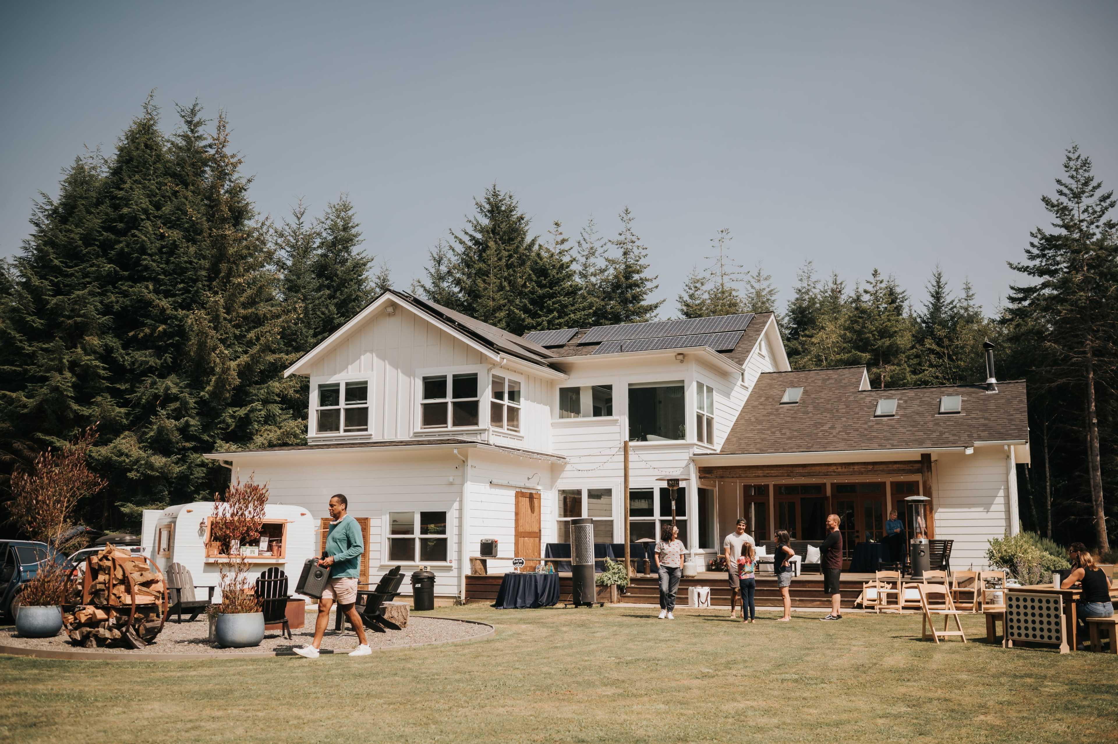 A group of people gather outside a large white house surrounded by trees, with some engaging in activities on the lawn.