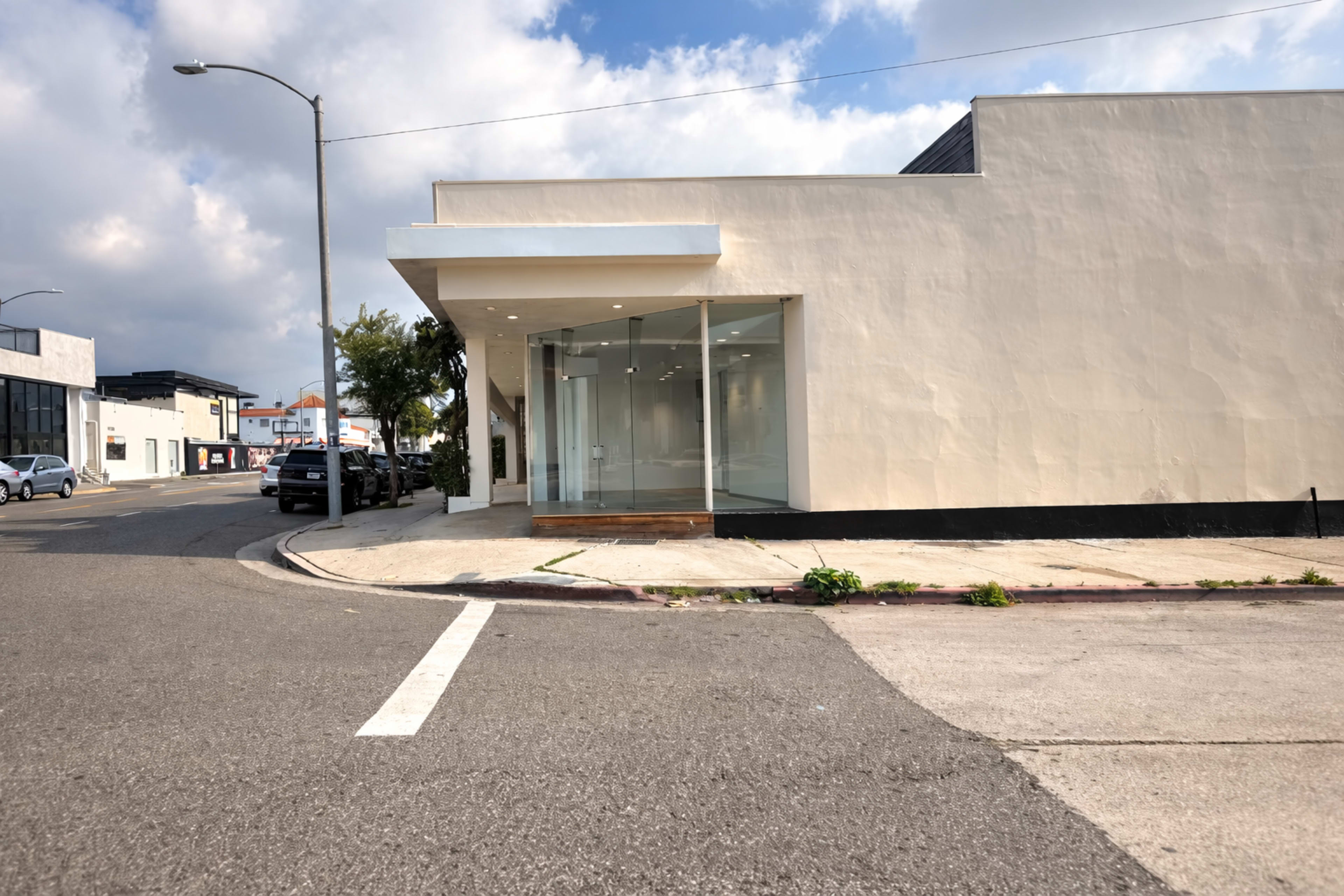 A modern, vacant storefront with large glass windows is located at the corner of a city street, surrounded by parked cars and a cloudy sky.