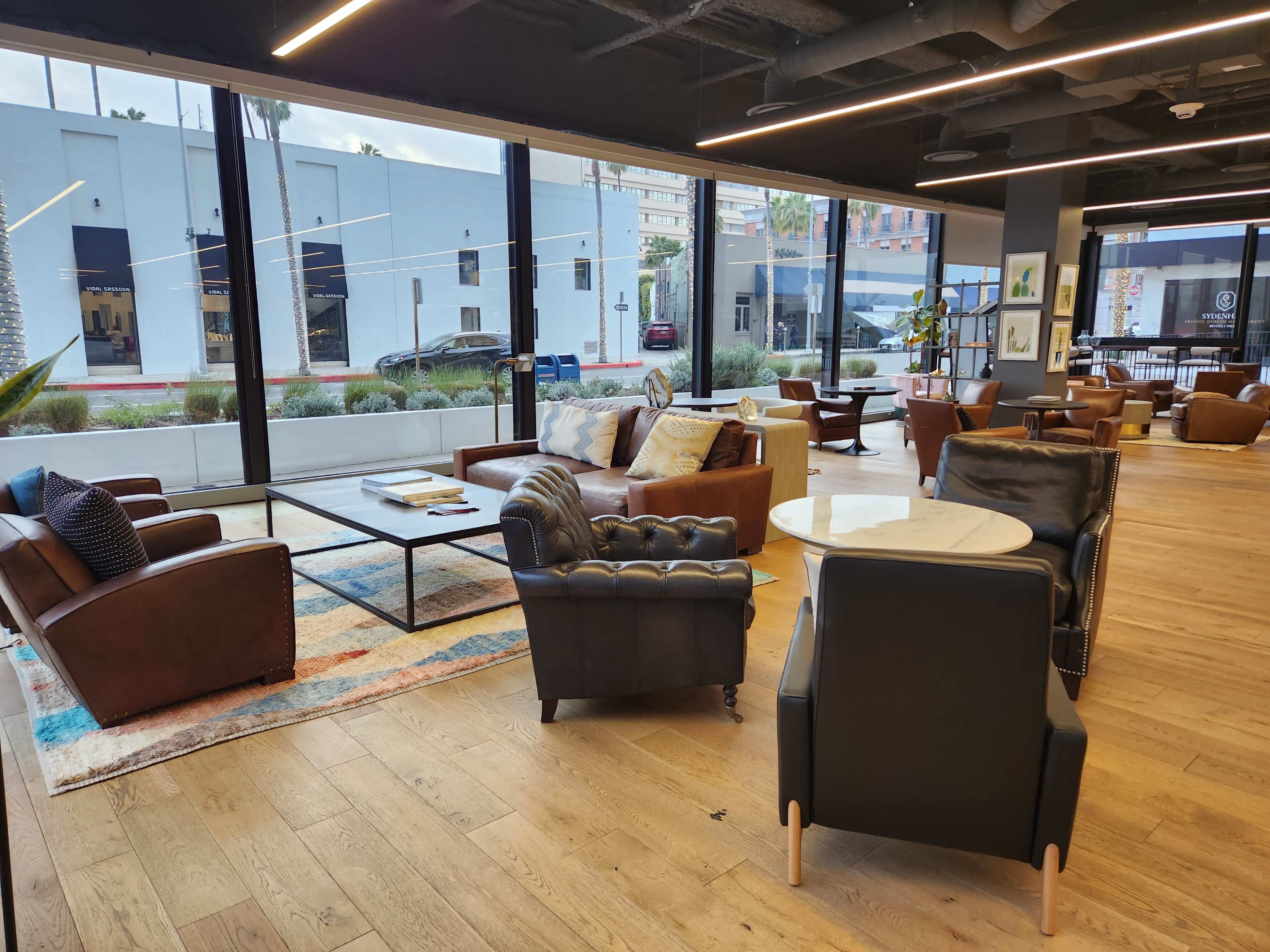 A cozy lounge area with large windows, featuring leather sofas, wooden floors, and a mix of tables and chairs.