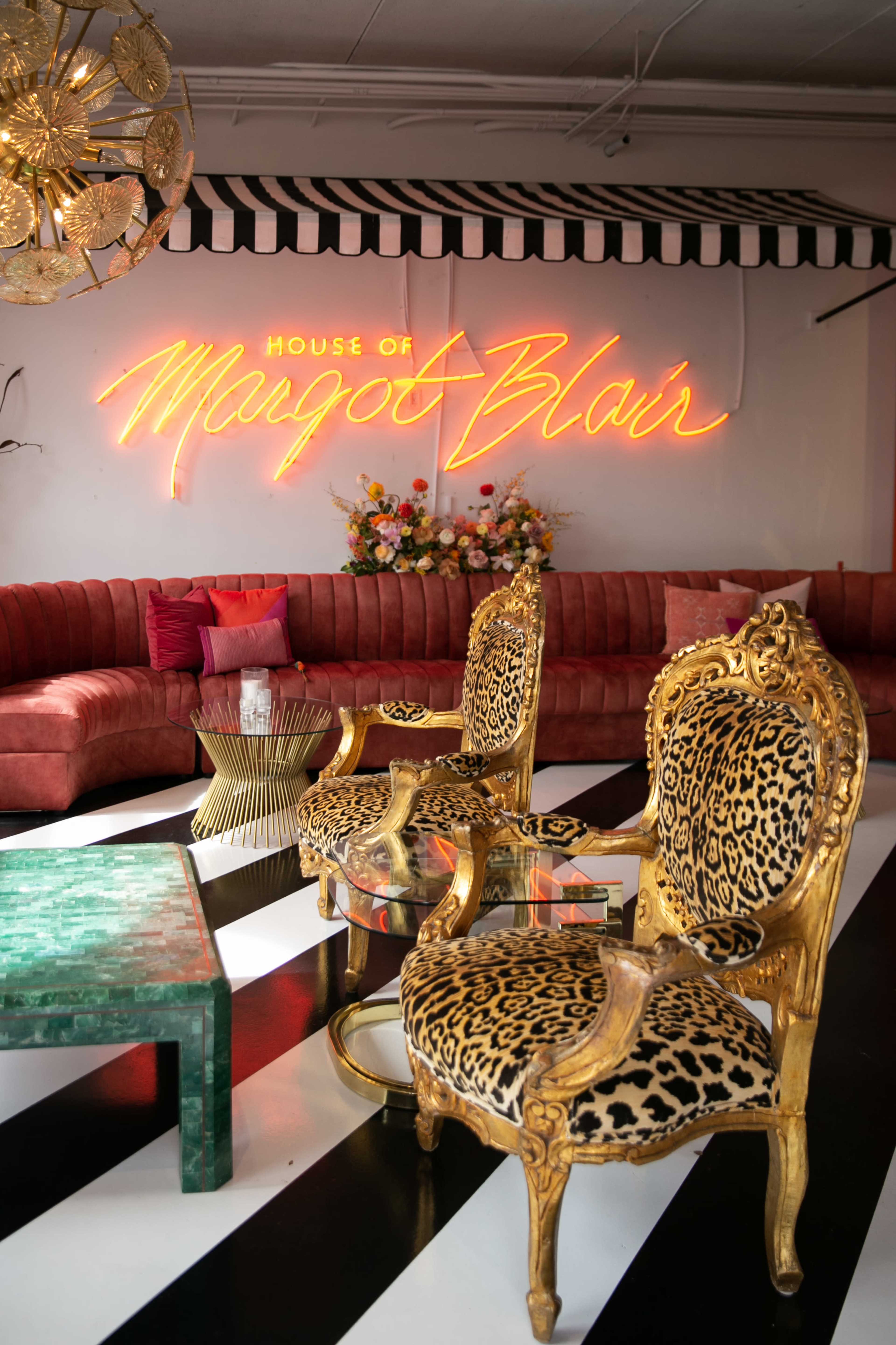 A stylish interior featuring two gold-framed chairs with leopard print fabric, a green table, and a neon sign that reads "House of Margot Blair" against a pink velvet couch backdrop.