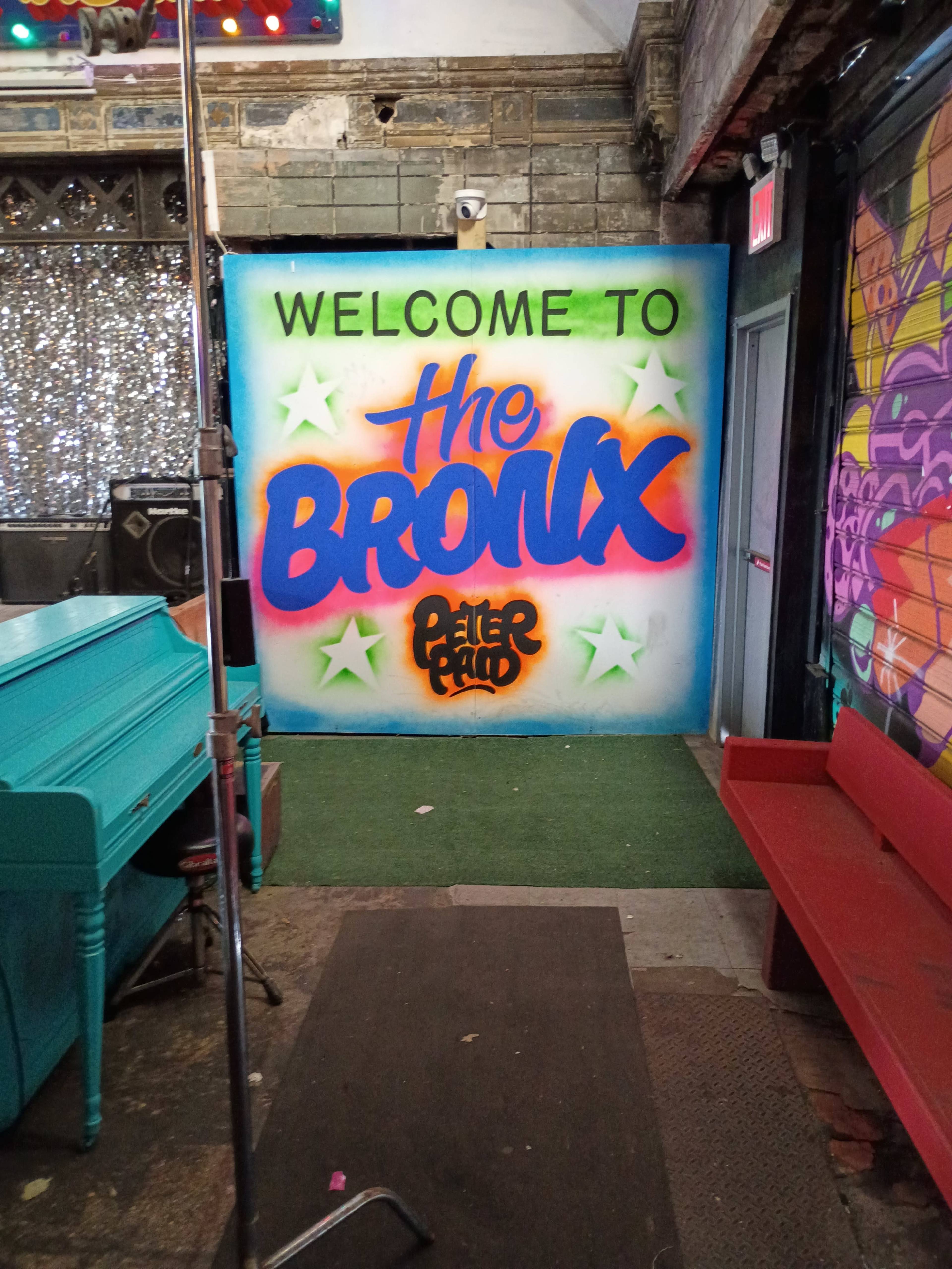 The image shows a vibrant mural that reads "WELCOME TO the BRONX" against a colorful background, situated in a venue with a turquoise piano and red seating.