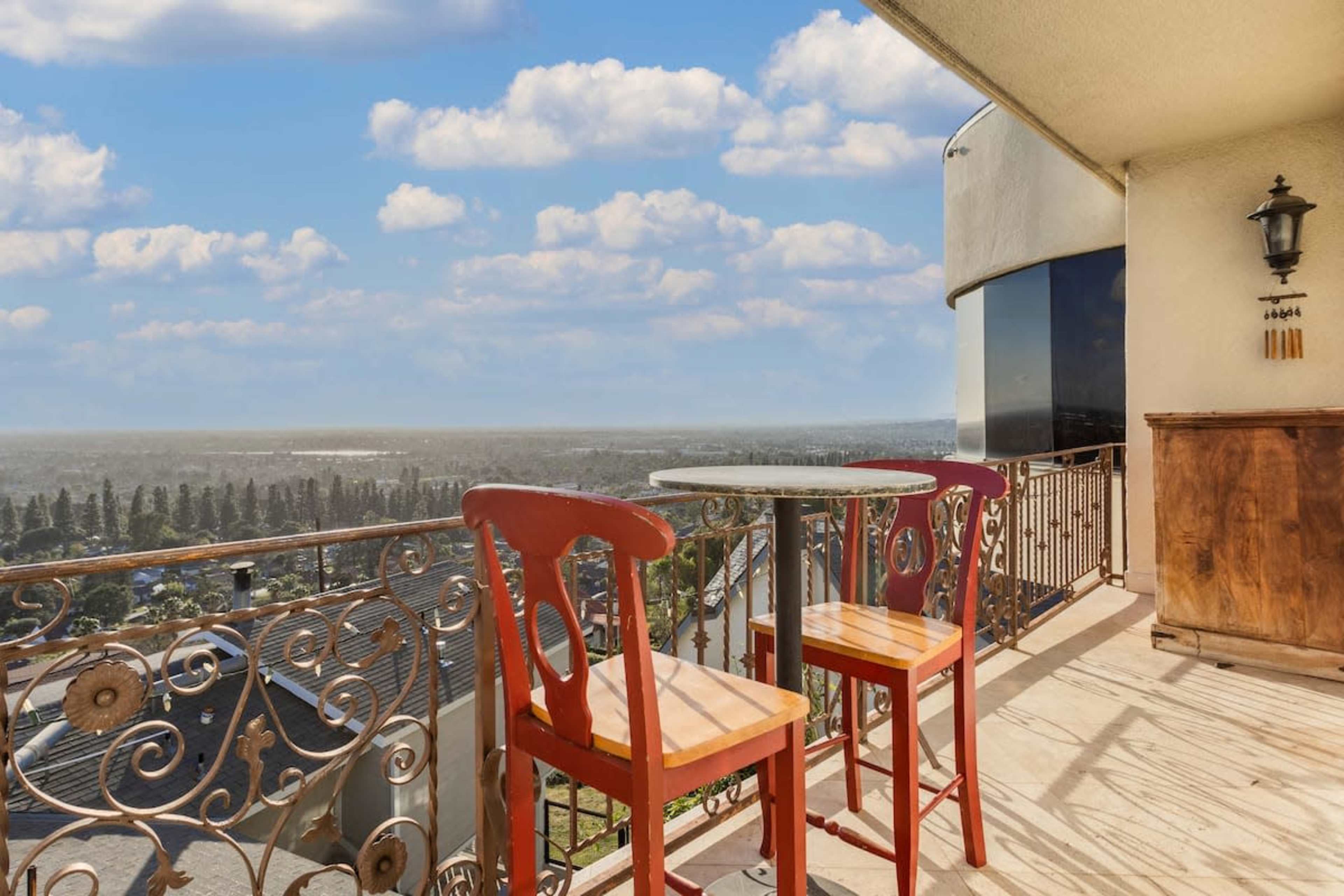 A balcony features a round table and two orange chairs overlooking a scenic view of the landscape below.