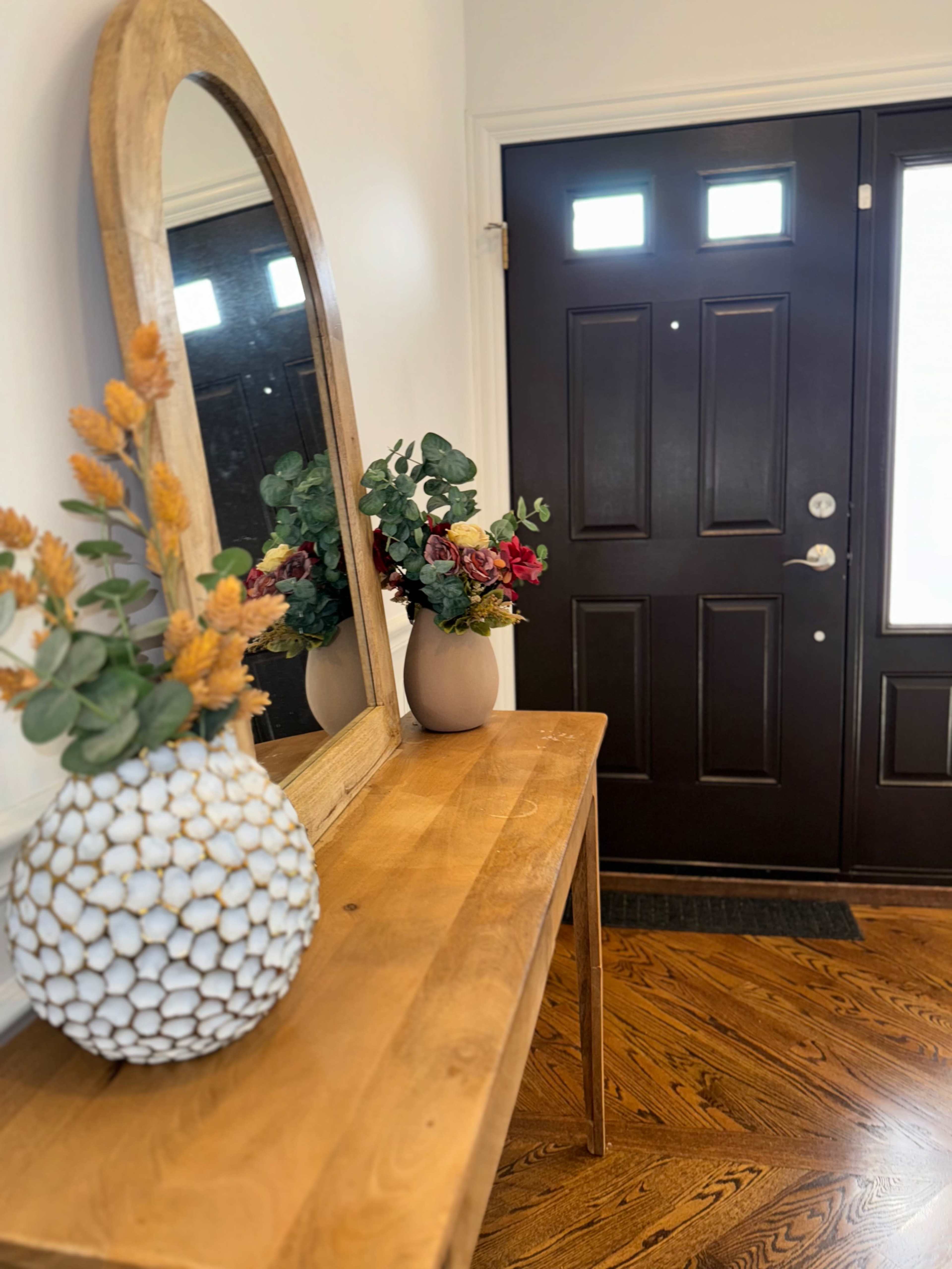 A wooden console table with decorative items and a mirror is positioned near a dark front door in an entryway.