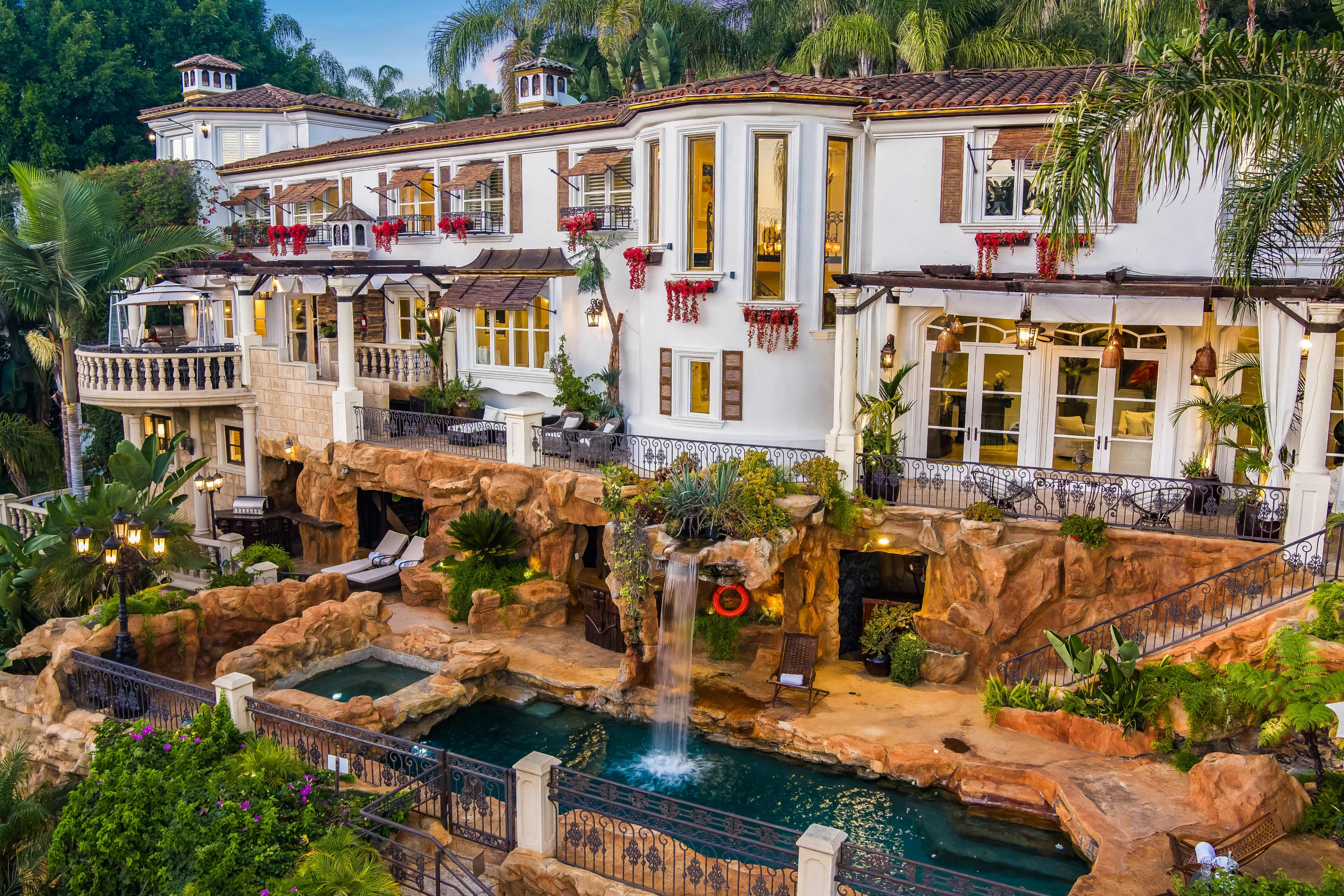 The image shows a large, multi-story mansion with a stone waterfall and a pool surrounded by lush greenery.