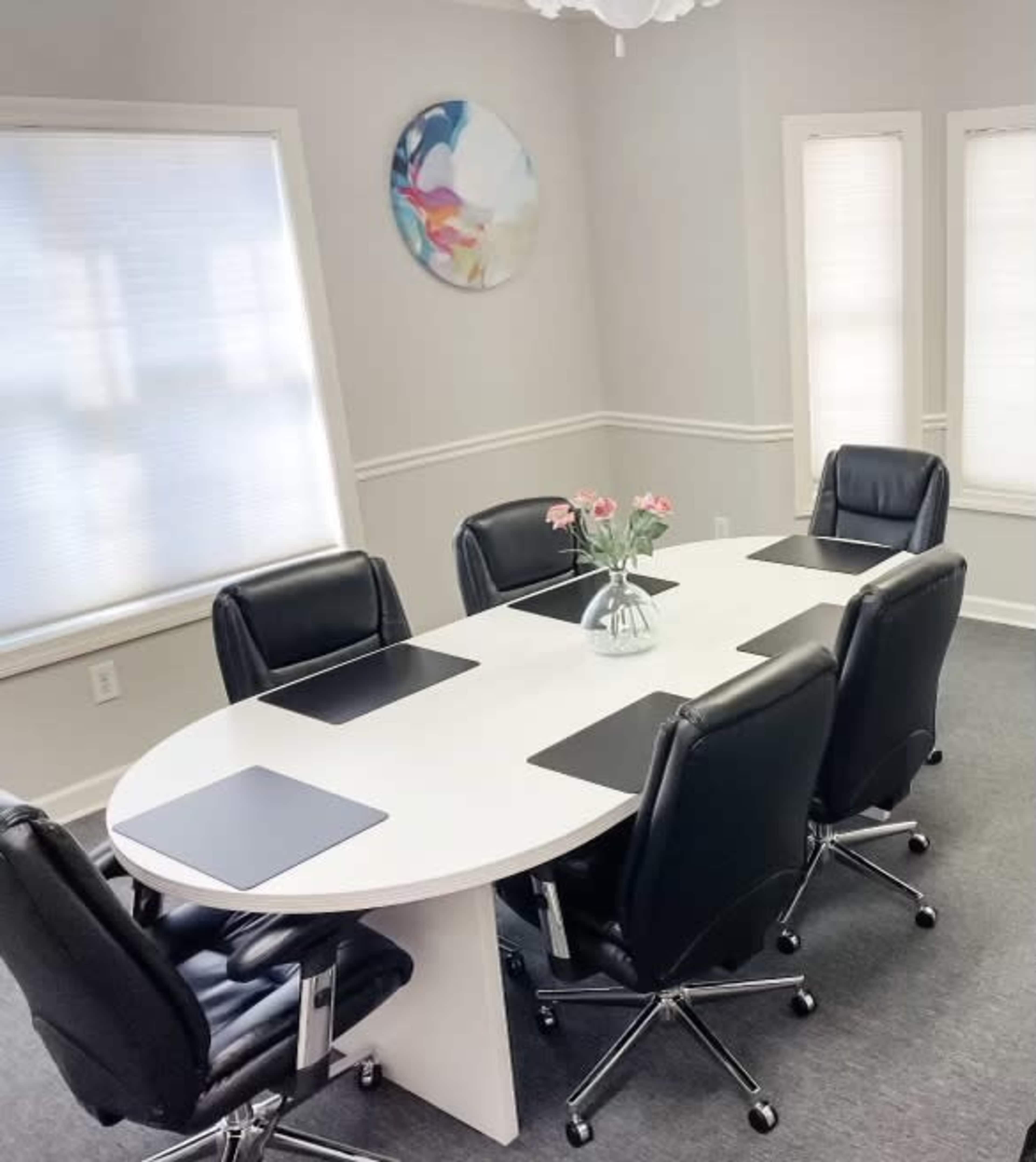 A modern conference room features an oval white table surrounded by six black office chairs, with a floral centerpiece and abstract artwork on the wall.