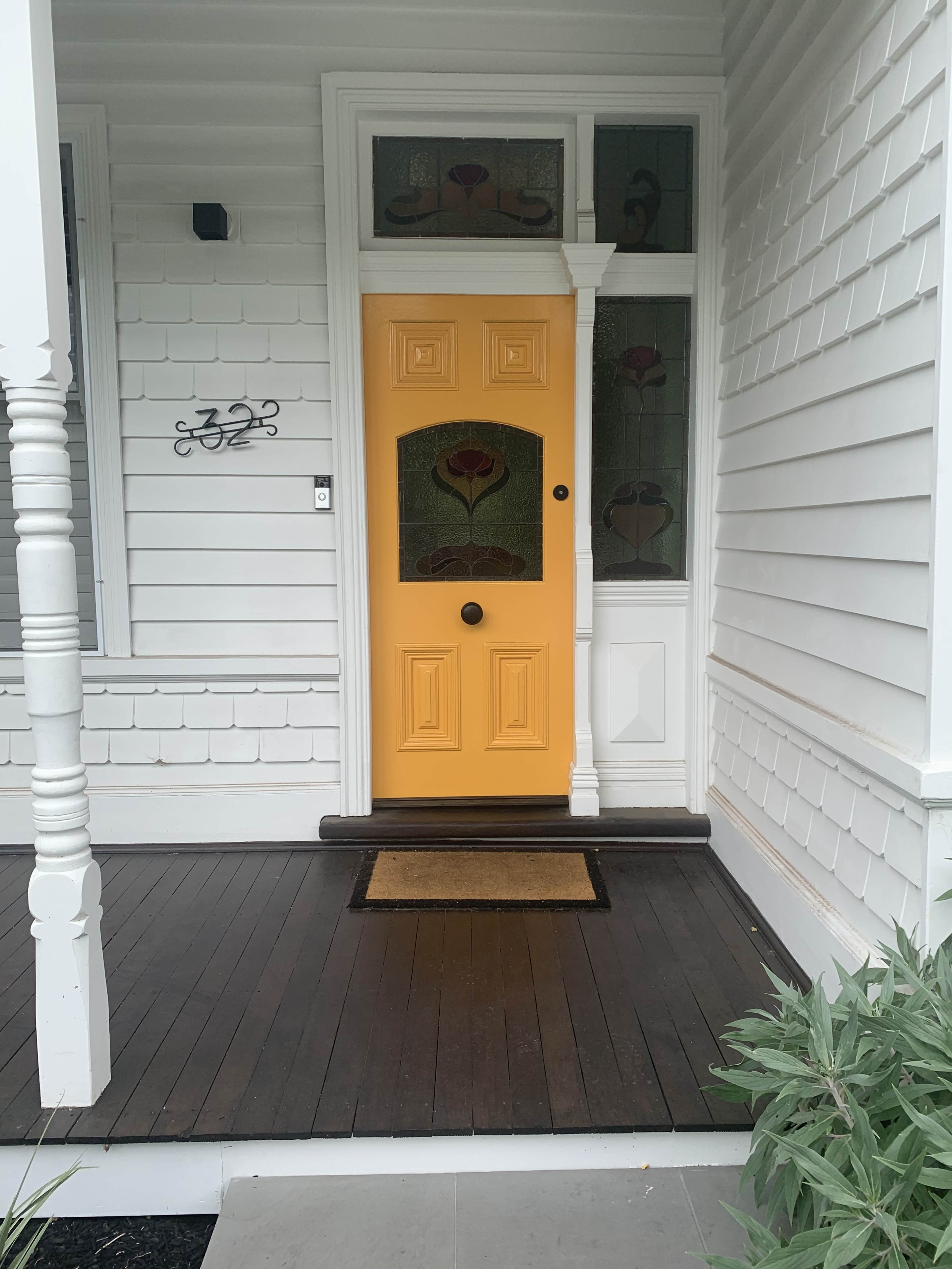 A yellow front door with decorative glass panels, located on a white house with a wooden porch.