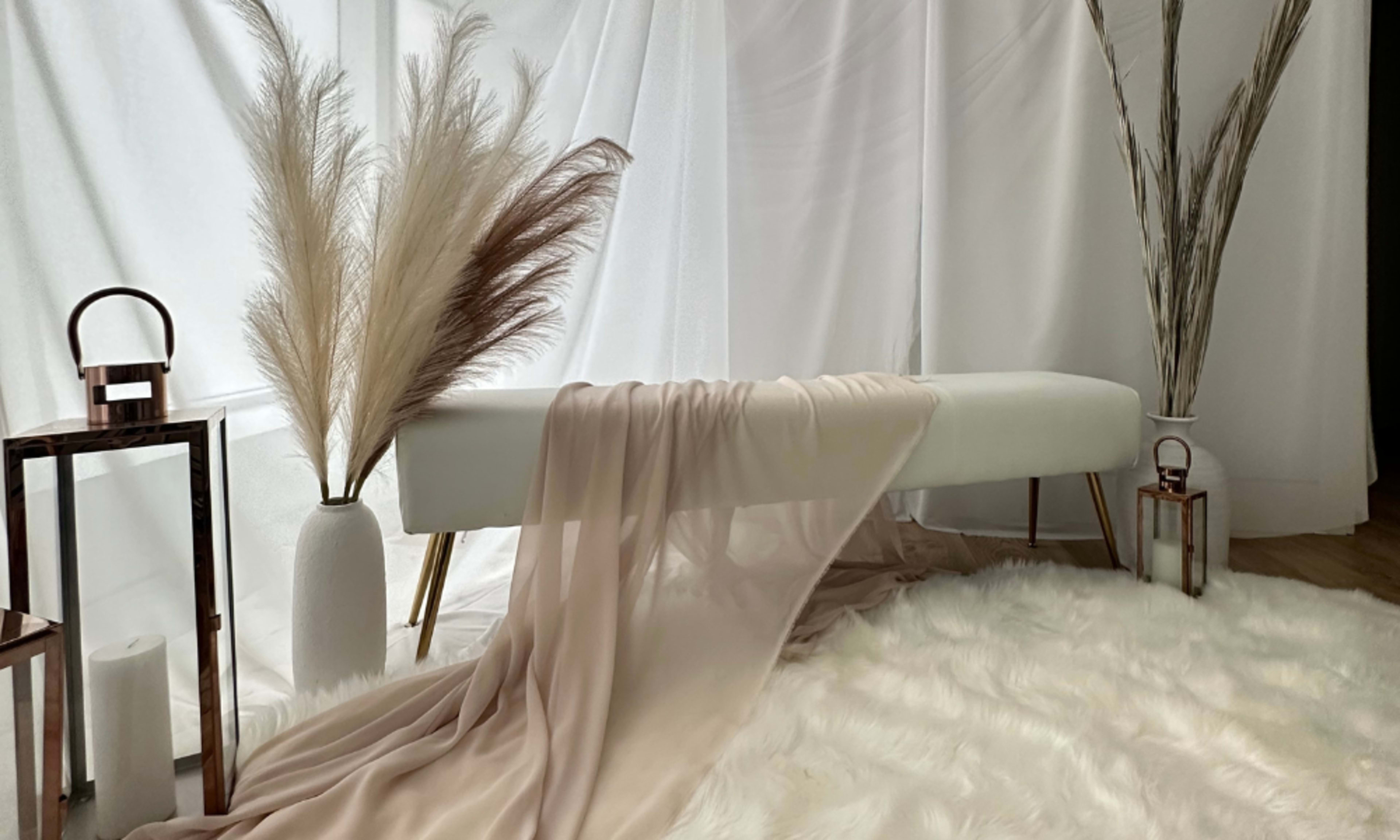 The scene features a white bench draped with a soft fabric, surrounded by decorative pampas grass and a cozy fur rug.