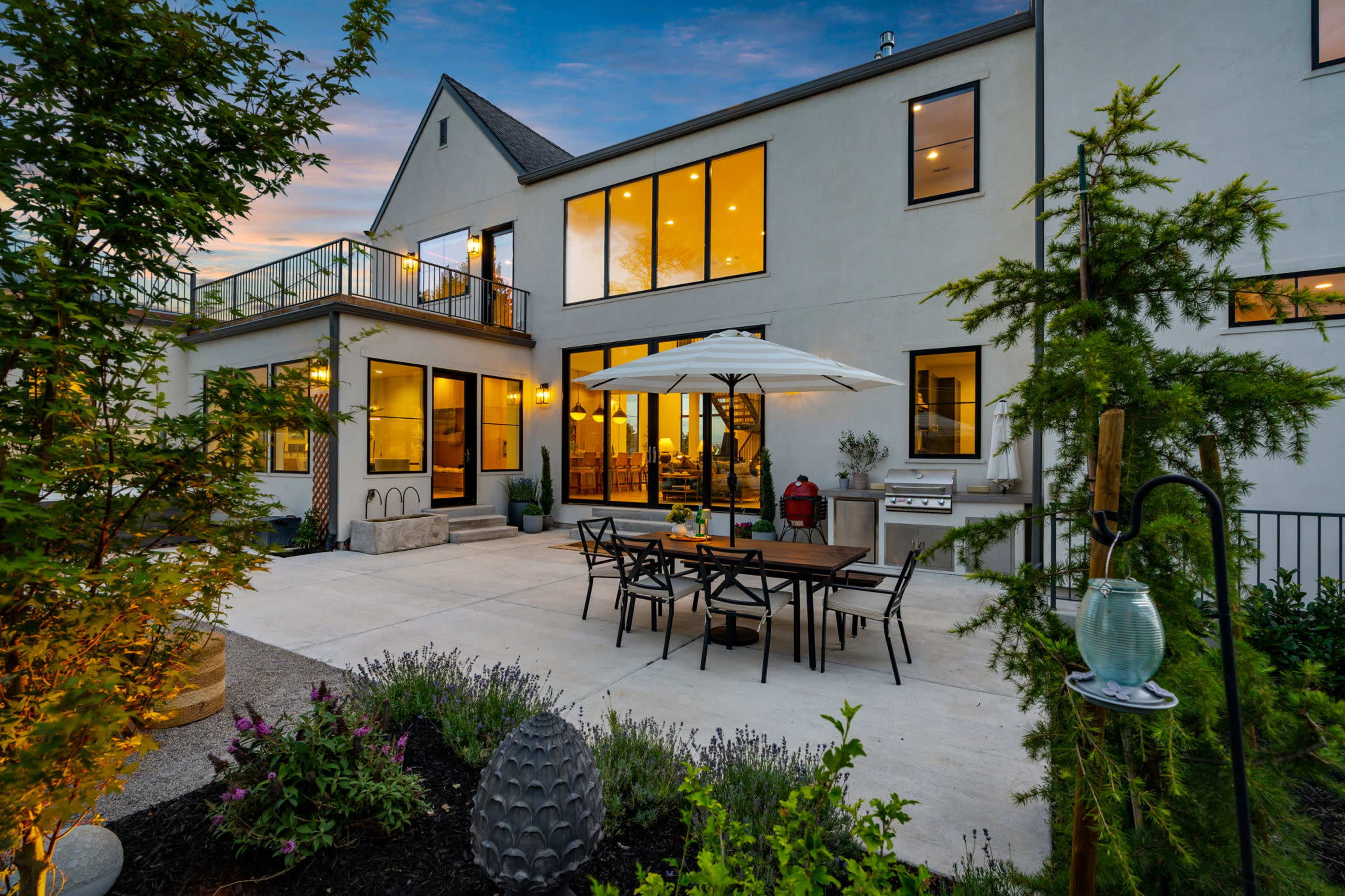 A modern patio features a dining set with an umbrella, surrounded by greenery and leading to large glass doors of a contemporary house.