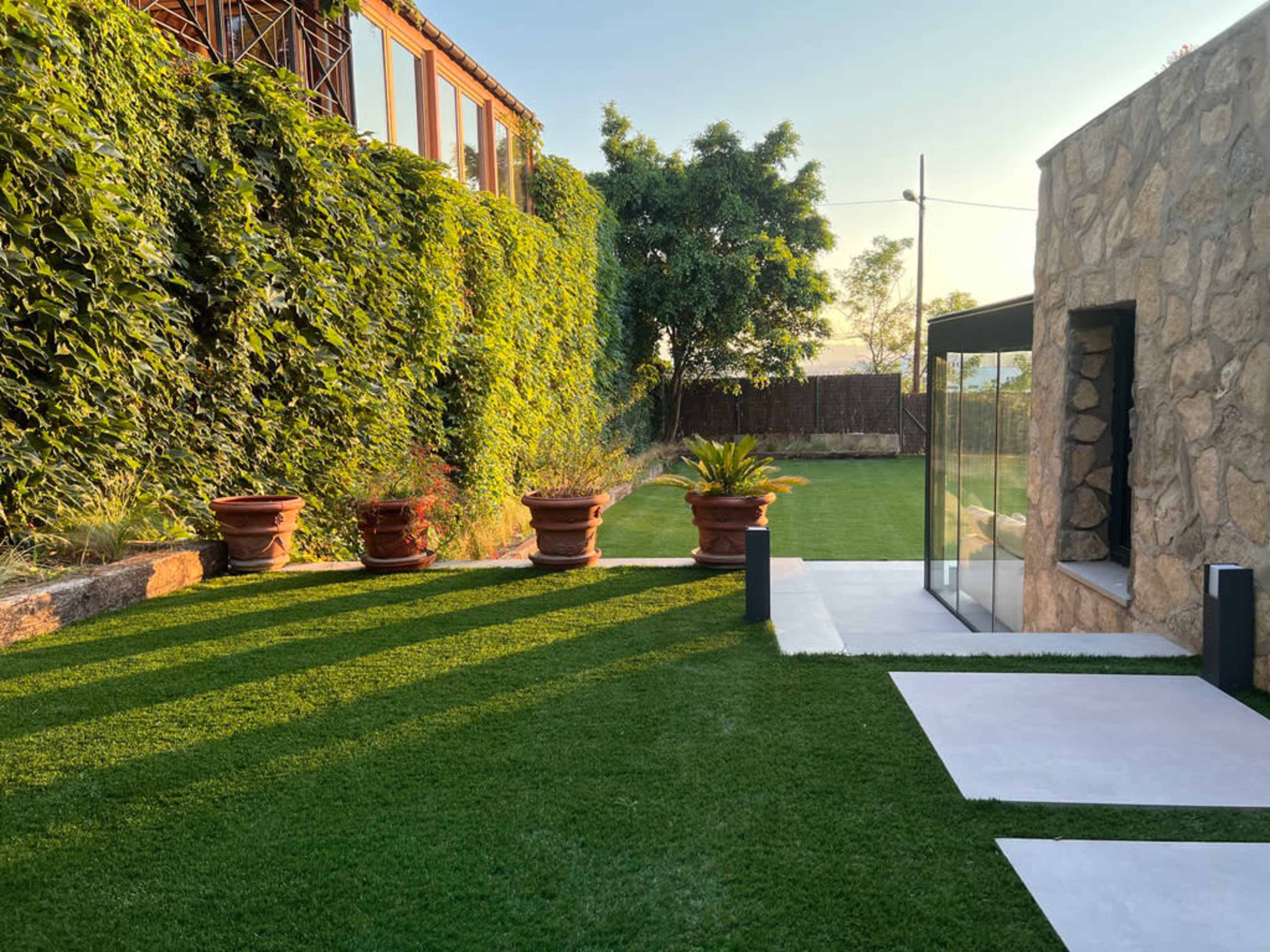 The image shows a landscaped garden with artificial grass, stone walls, potted plants, and a modern structure in the background.