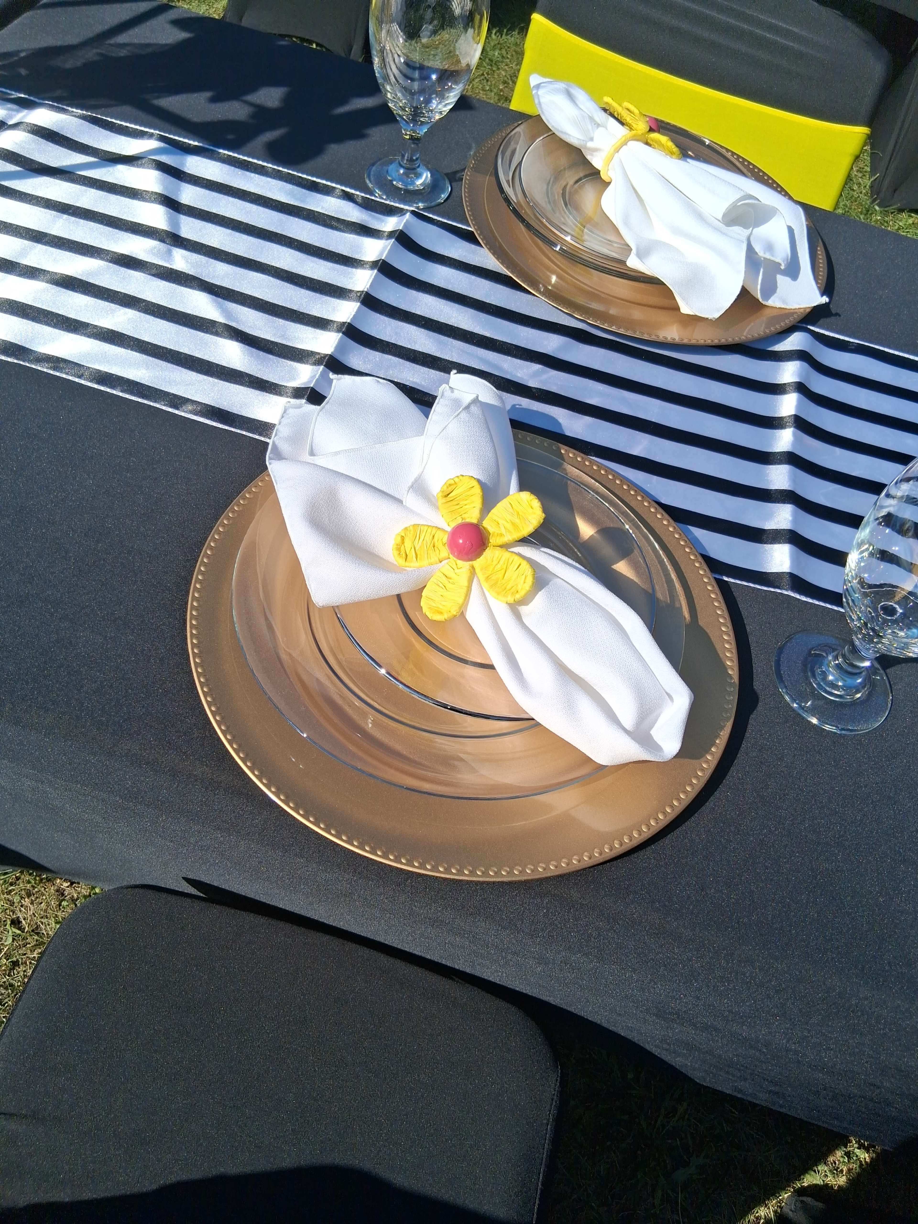 The image shows a dining table set with black and white striped table runners, gold plates, and neatly folded white napkins adorned with yellow flower accents.