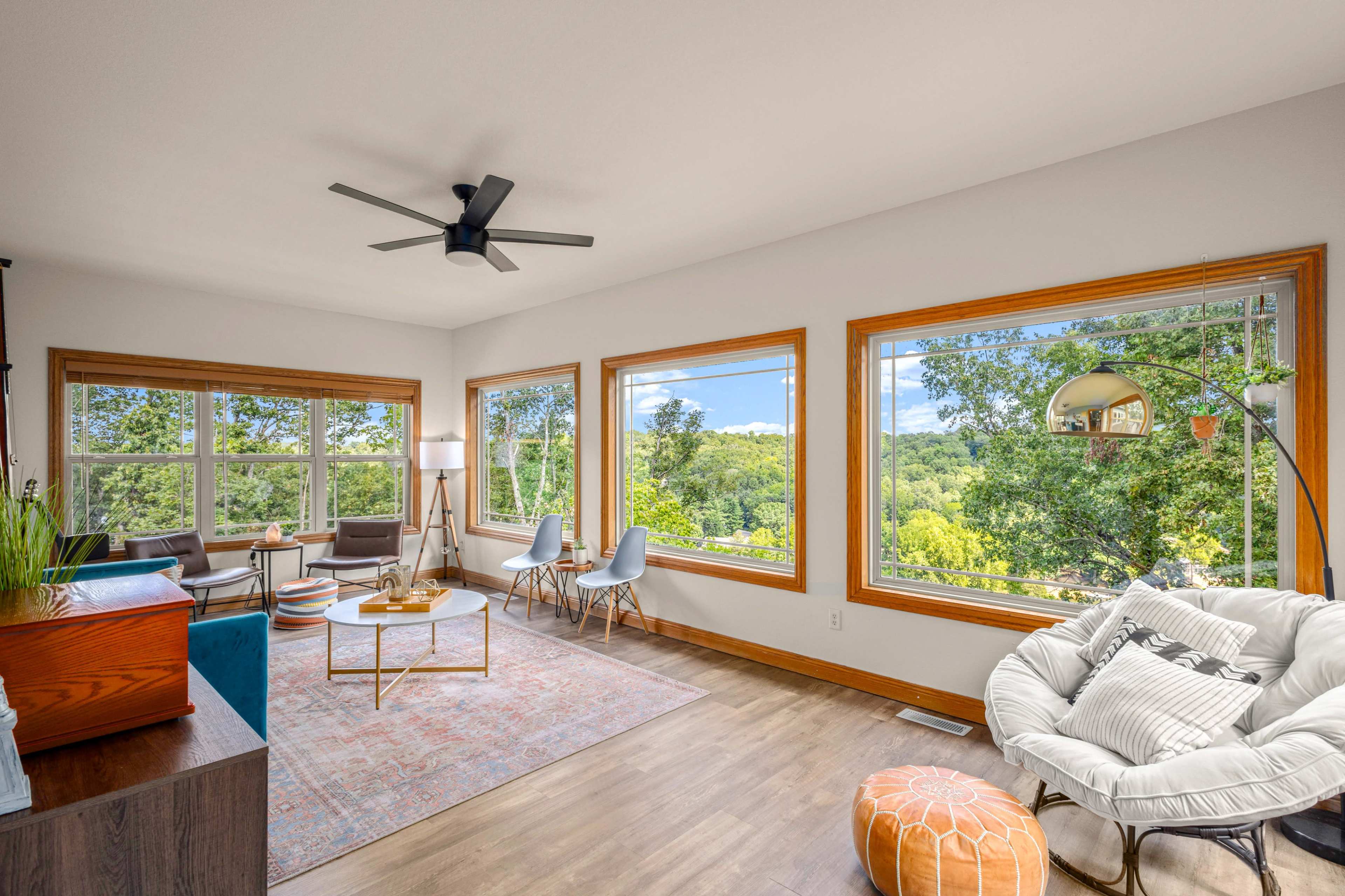 A living room features large windows with a view of greenery, a round coffee table, modern furniture, and a ceiling fan.