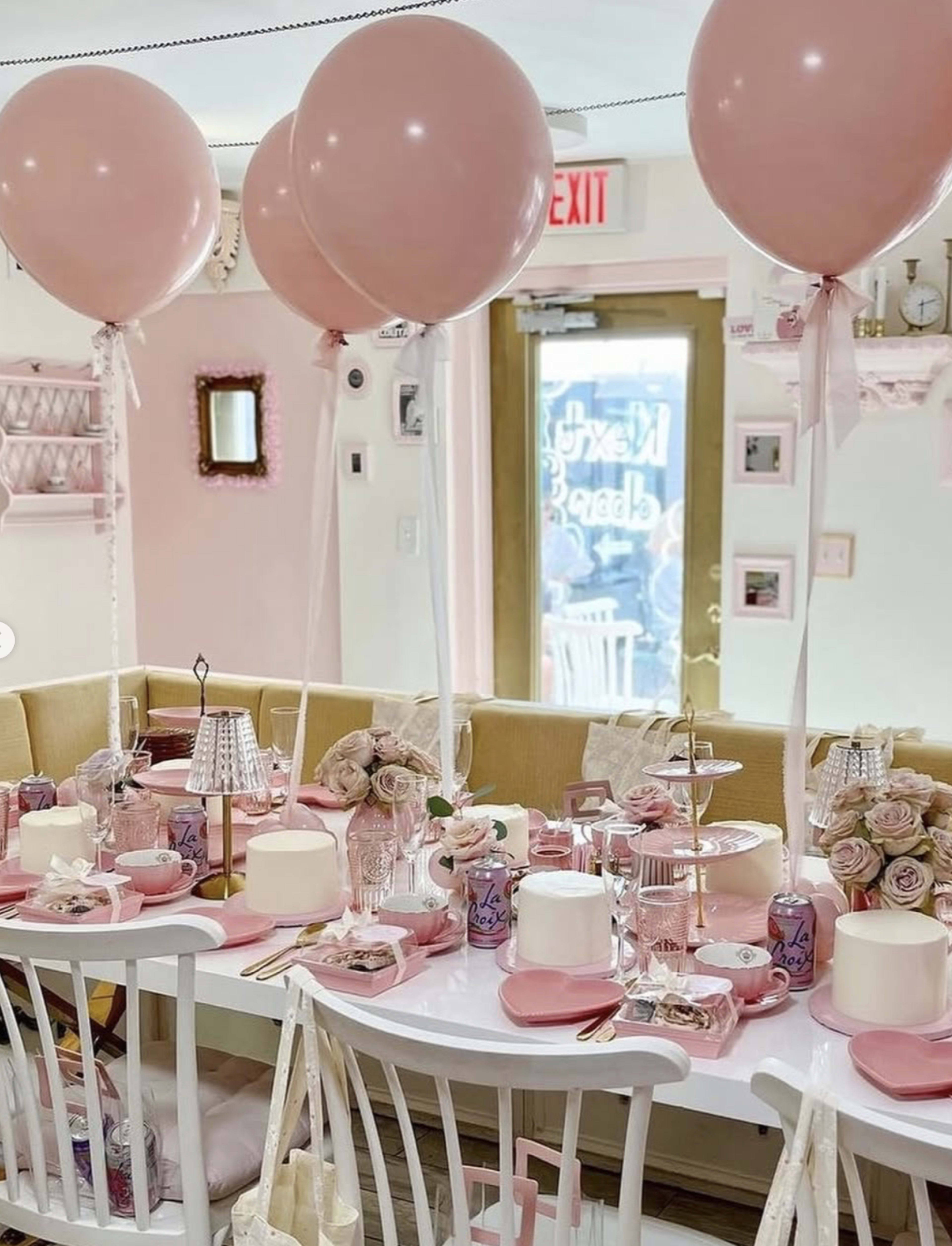A table is elegantly set with pink decorations, including balloons, flowers, and fine dining ware, in a bright room featuring pastel walls and a cozy layout.
