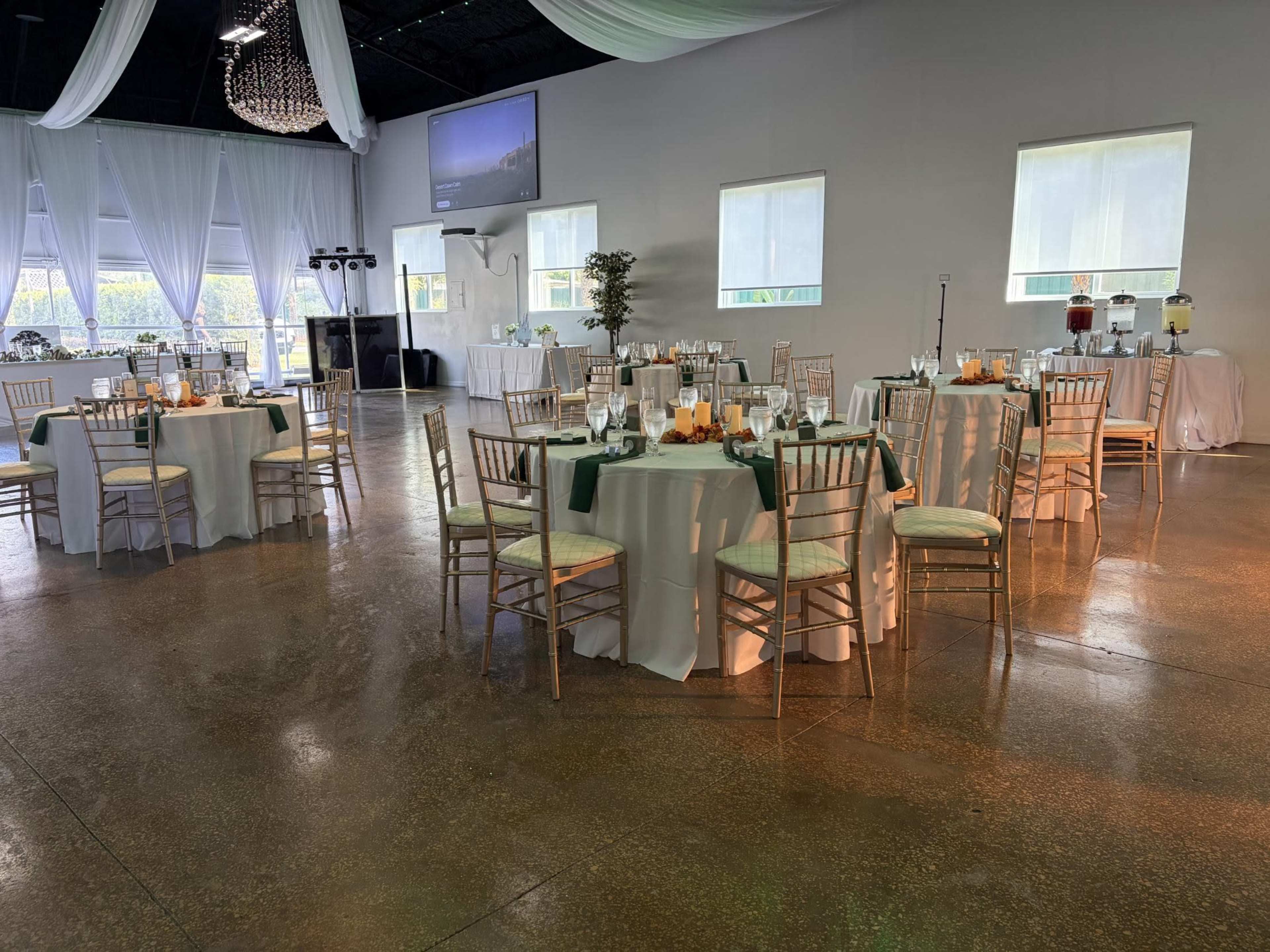 A spacious event hall features elegantly set tables with white tablecloths and gold chiavari chairs, surrounded by draped curtains and ambient lighting.