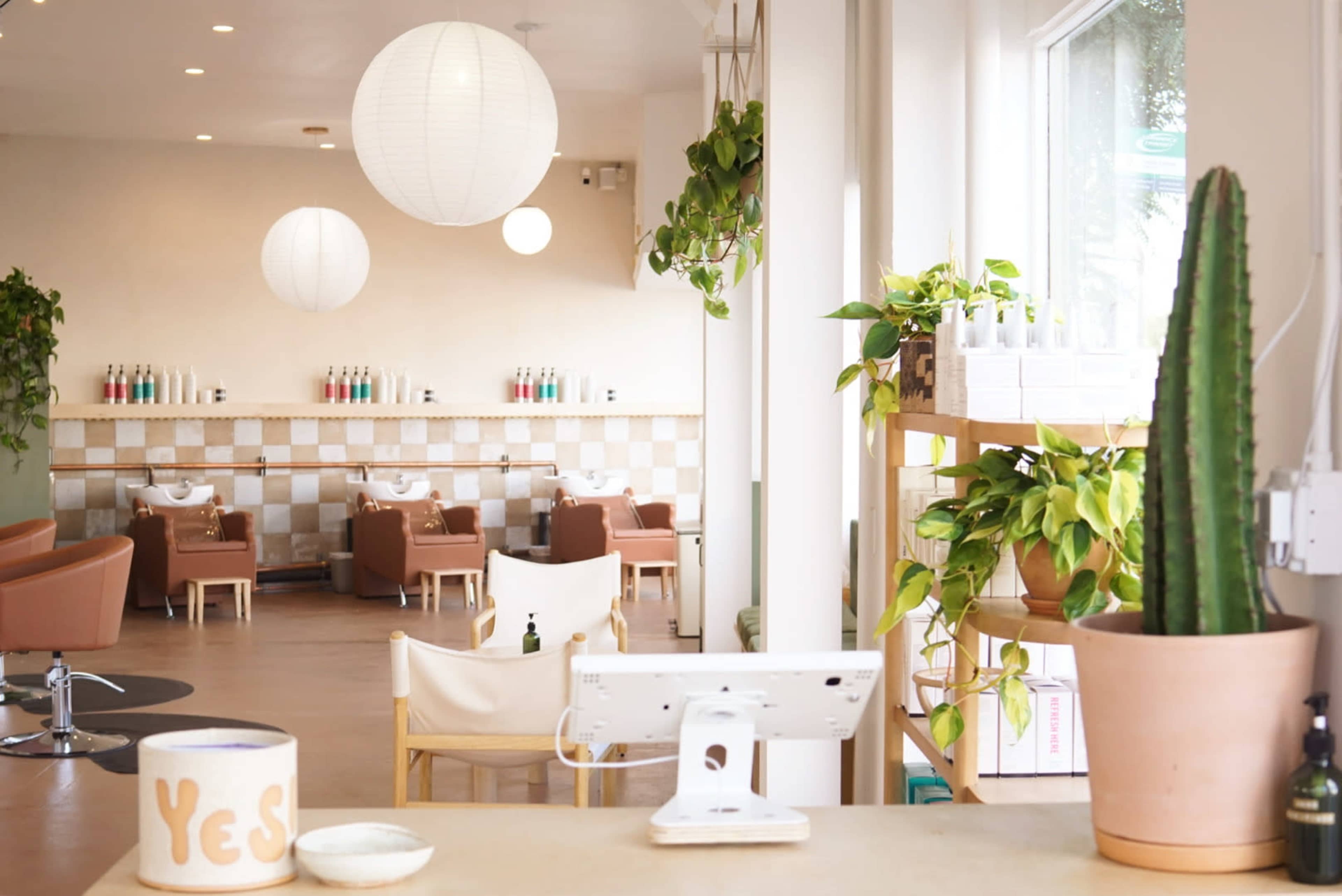 The image shows a bright and modern salon interior with hanging paper lanterns, green plants, and a reception area featuring a computer and decorative items.