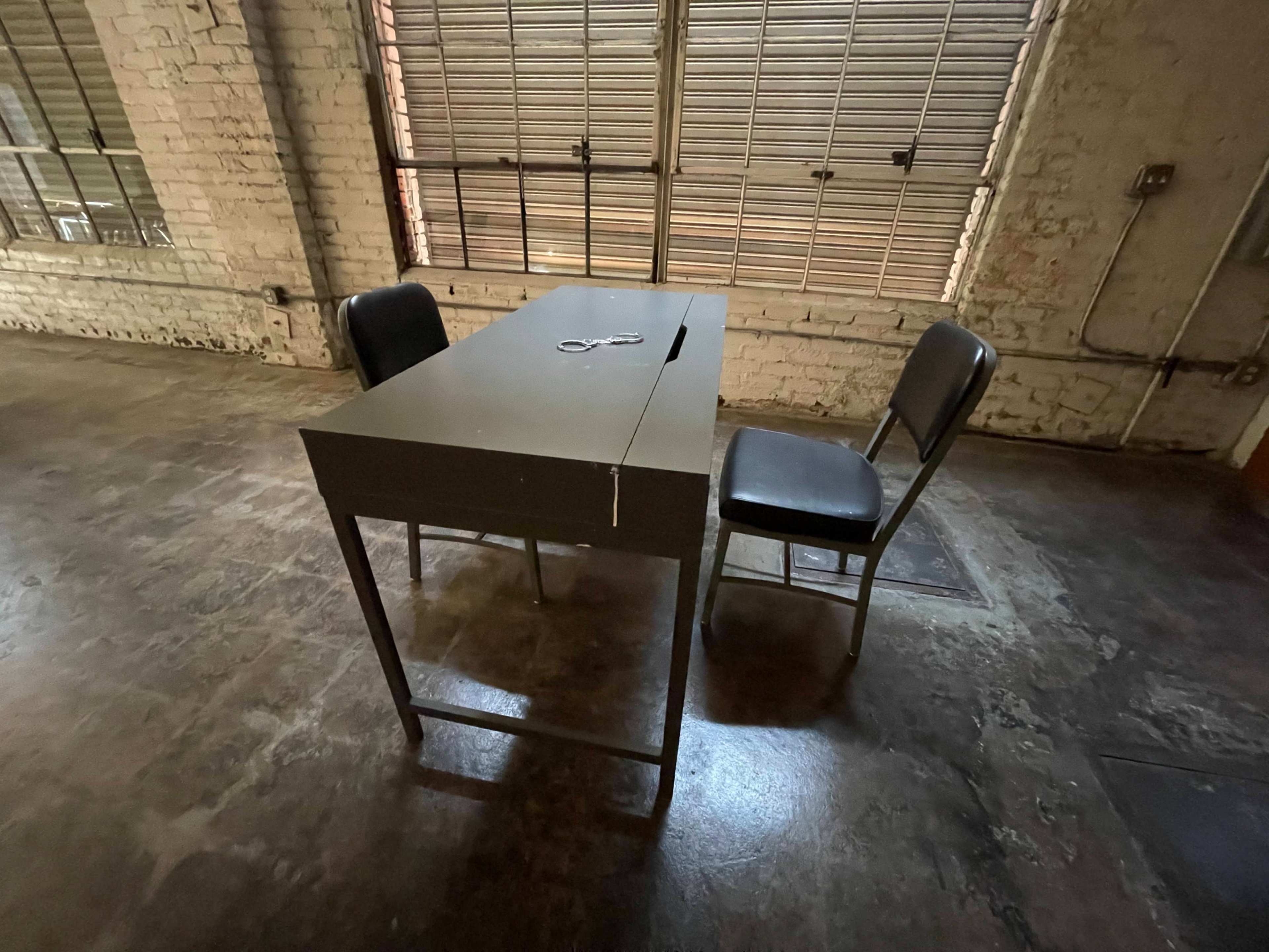 A bare metal table with a slit in the center and two black chairs sit on a concrete floor beneath large windows with metal grilles.