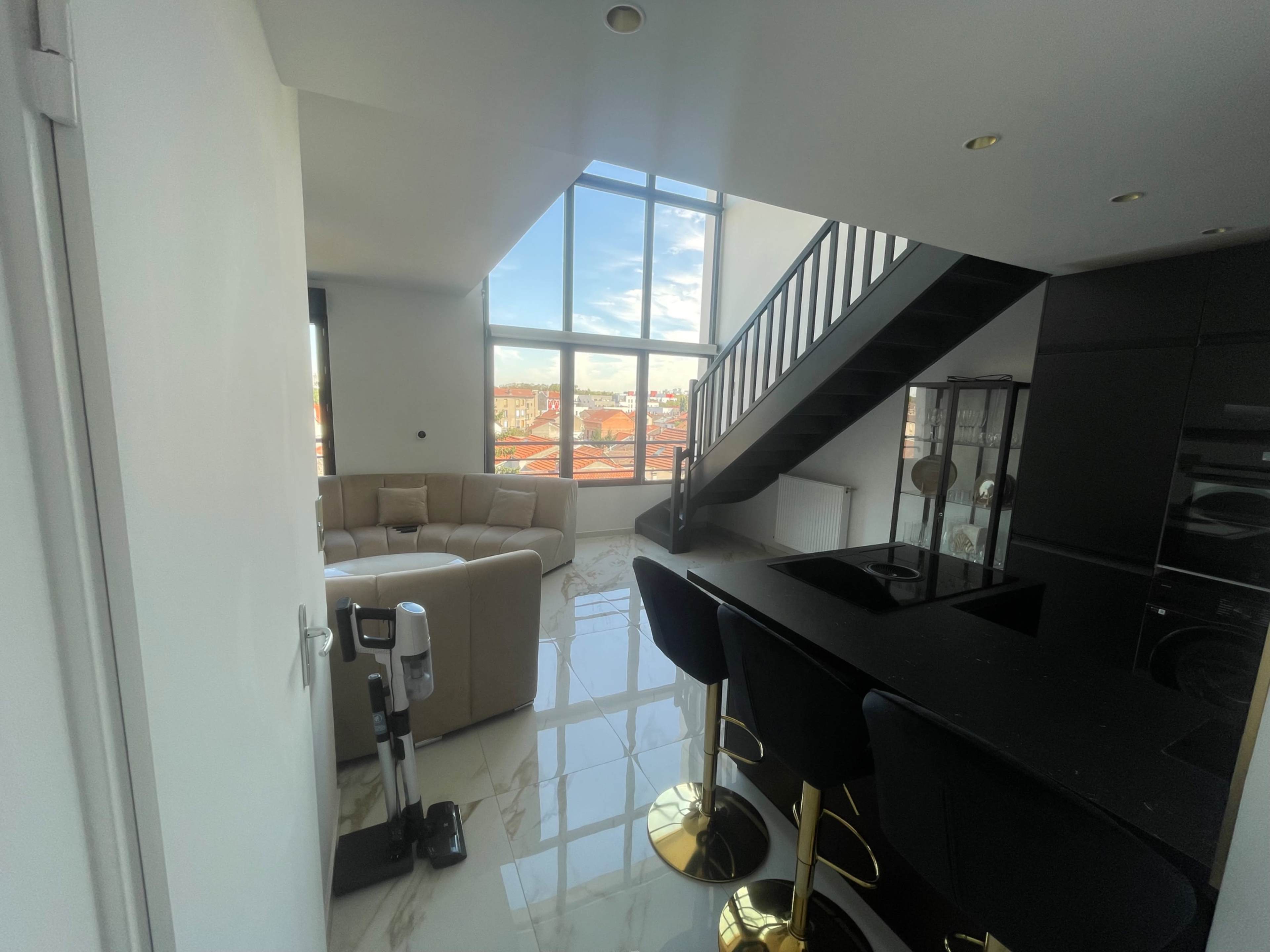 The image shows a modern interior space featuring a living area with a beige sectional sofa, a staircase, and a dining bar with black stools, all illuminated by large windows showcasing a city view.