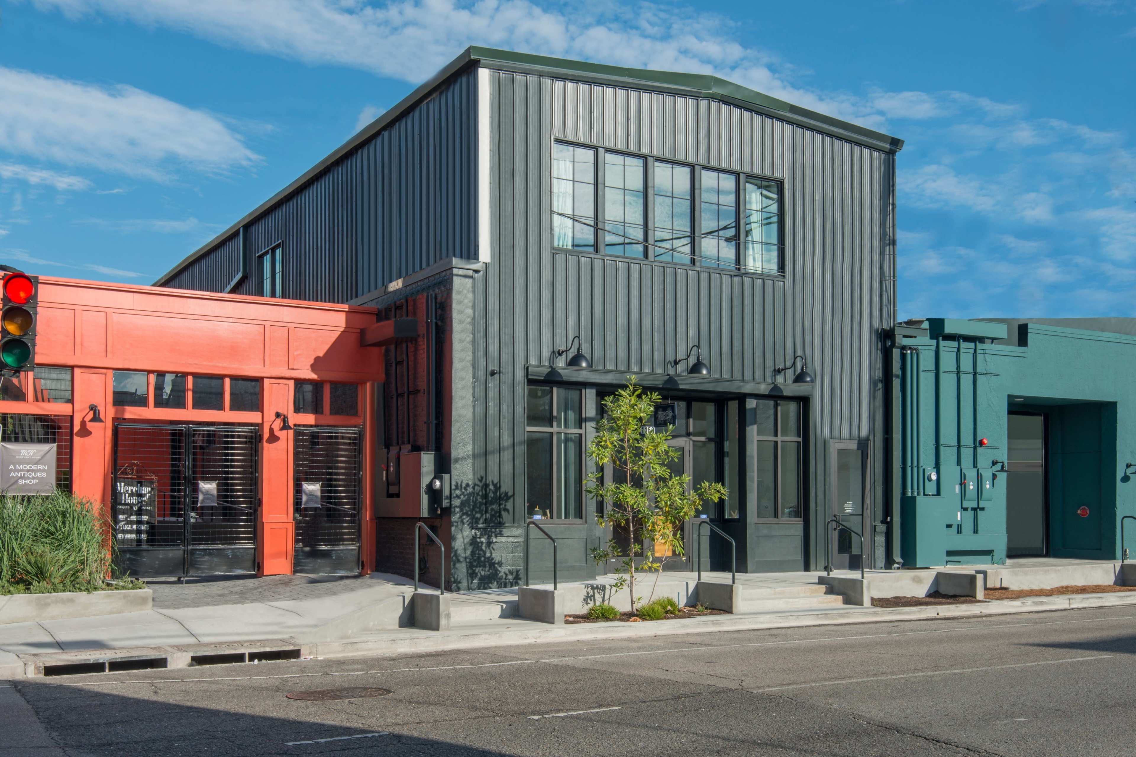 The image shows a modern building with a combination of red and dark green metal siding, featuring large windows and a small tree near the entrance.